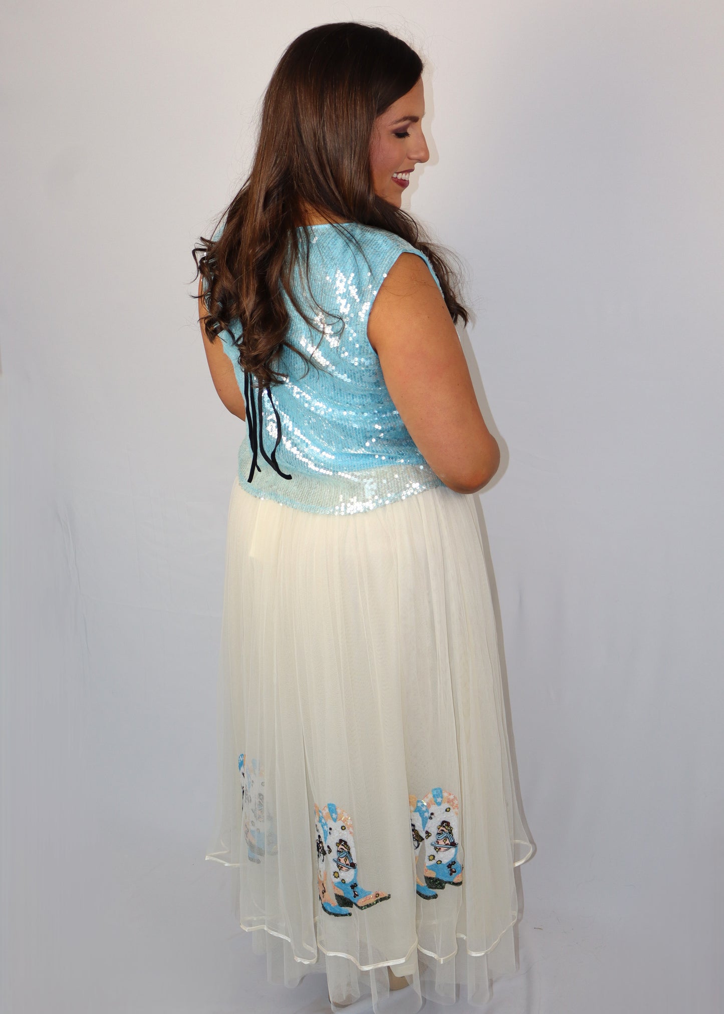 Woman wearing a blue sequin top and cream skirt with sequin cowboy boots embroidery on a plain background
