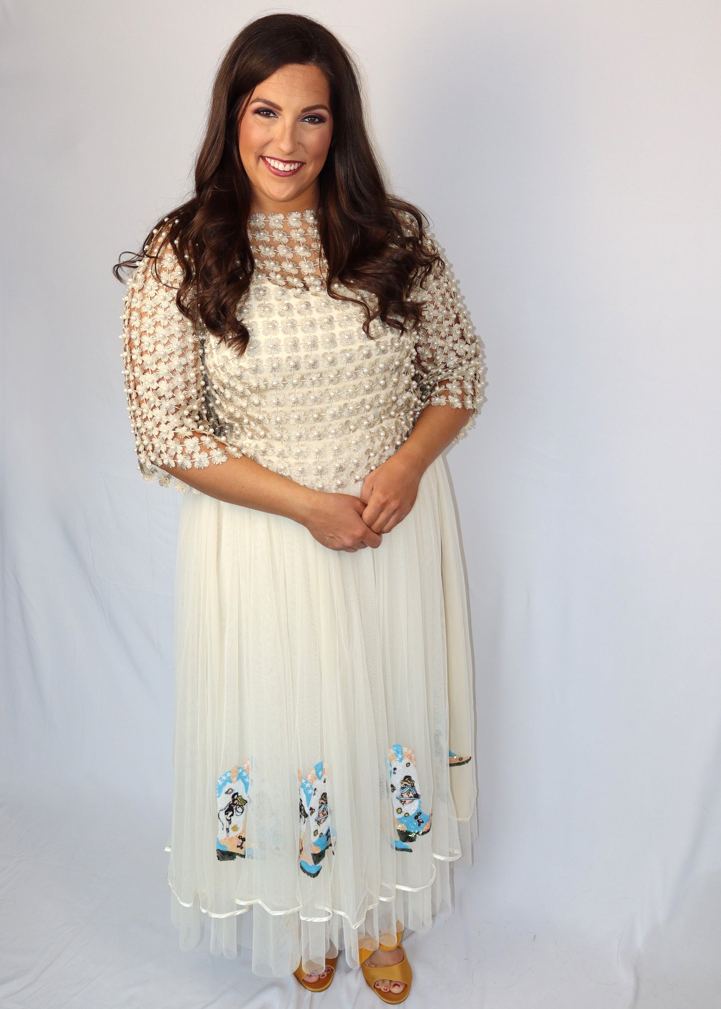 Woman wearing a gold pearl and rope-like top paired with a cream tulle skirt with sequin boots on it. She is standing against a plain, white background.
