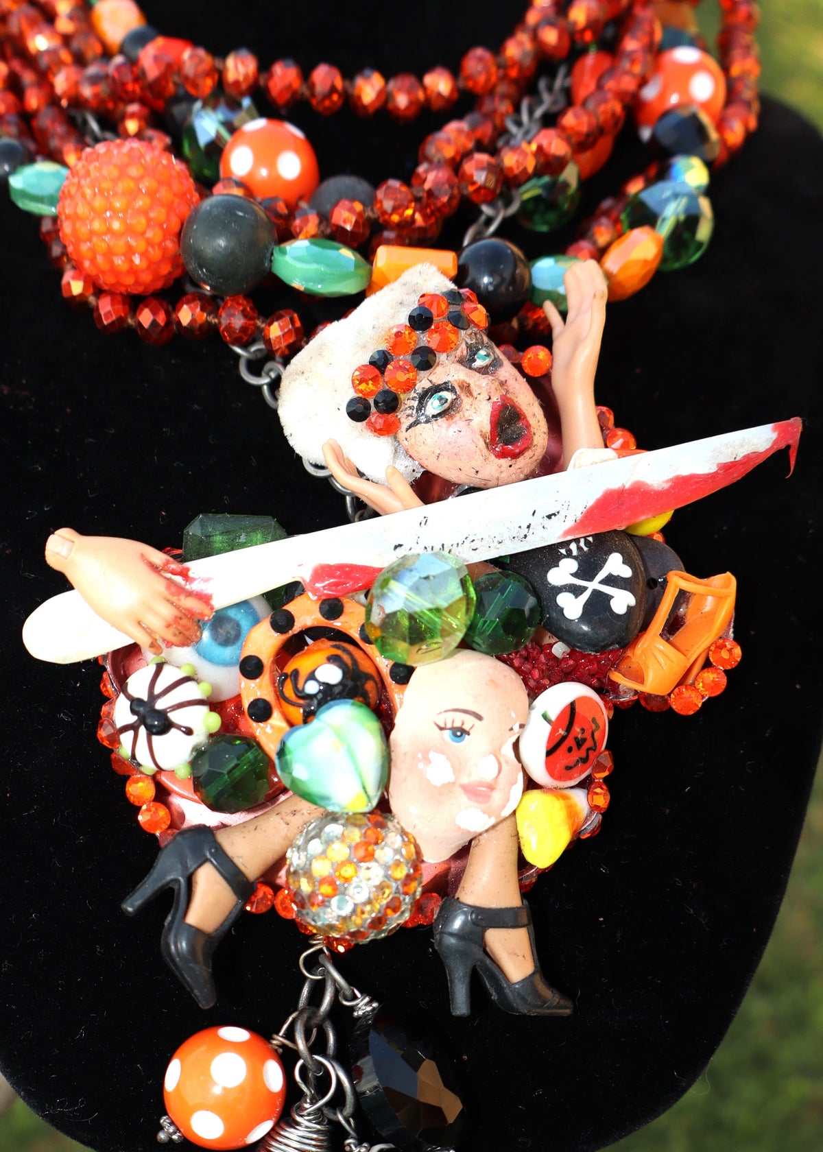 Orange, green, and black multi-strand beaded Art by Amy upcycled necklace with Halloween, scary movie-themed charms on a black stand against a grassy background