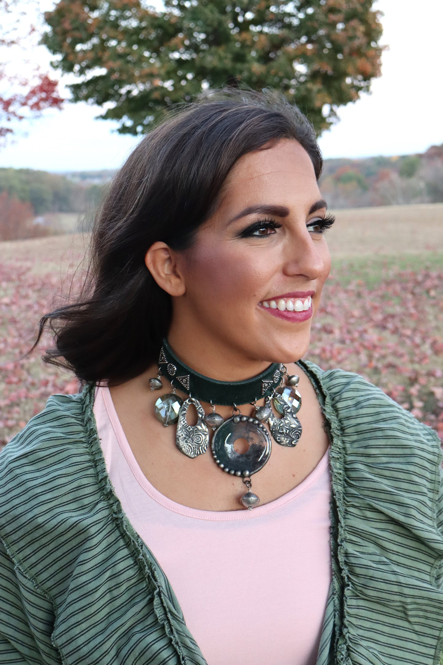 Woman wearing a dark green, leather choker necklace with a statement pendant and various other pendants in an outdoor setting