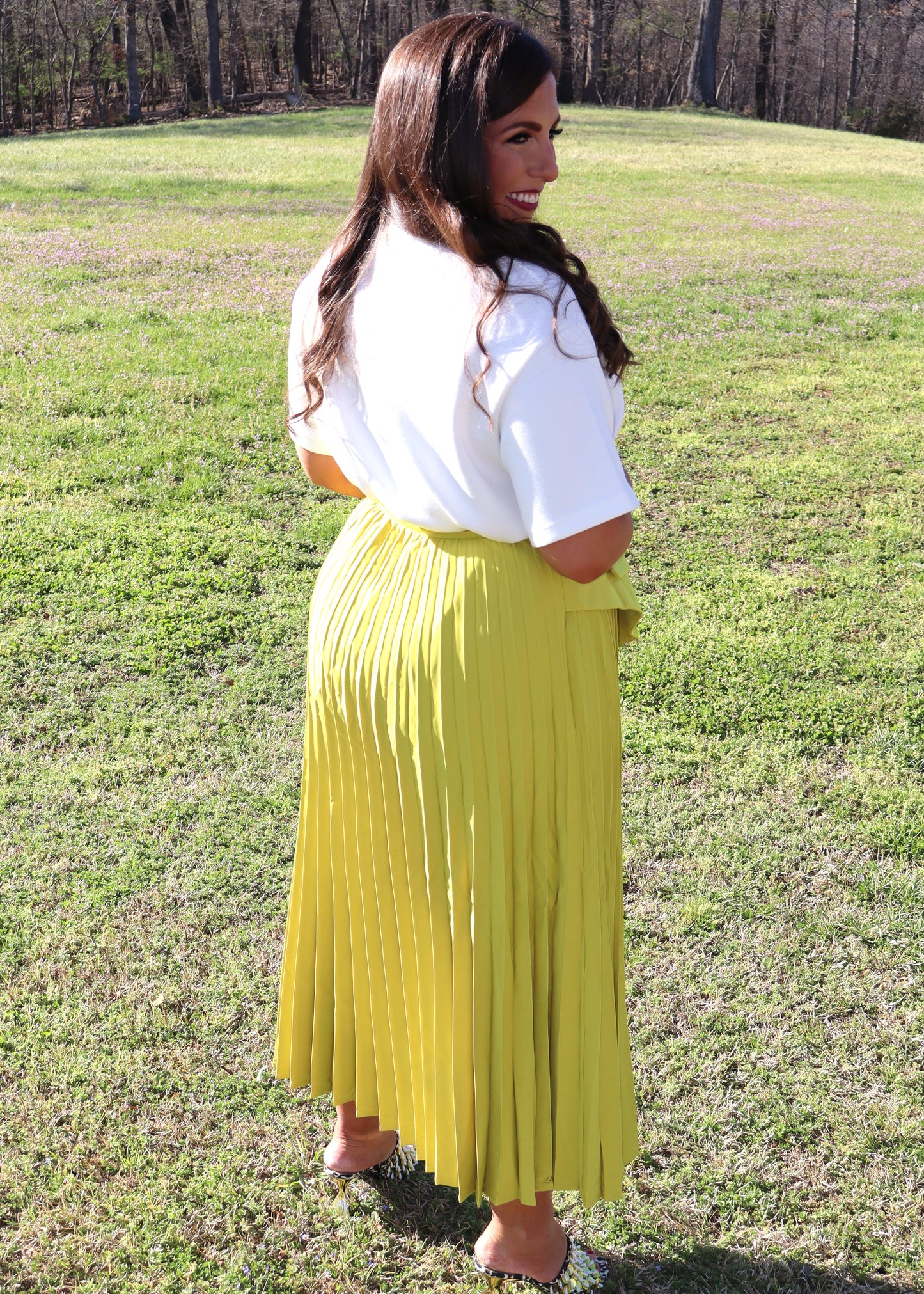 Woman wearing a white top and yellow pleated skirt standing in a grassy field.