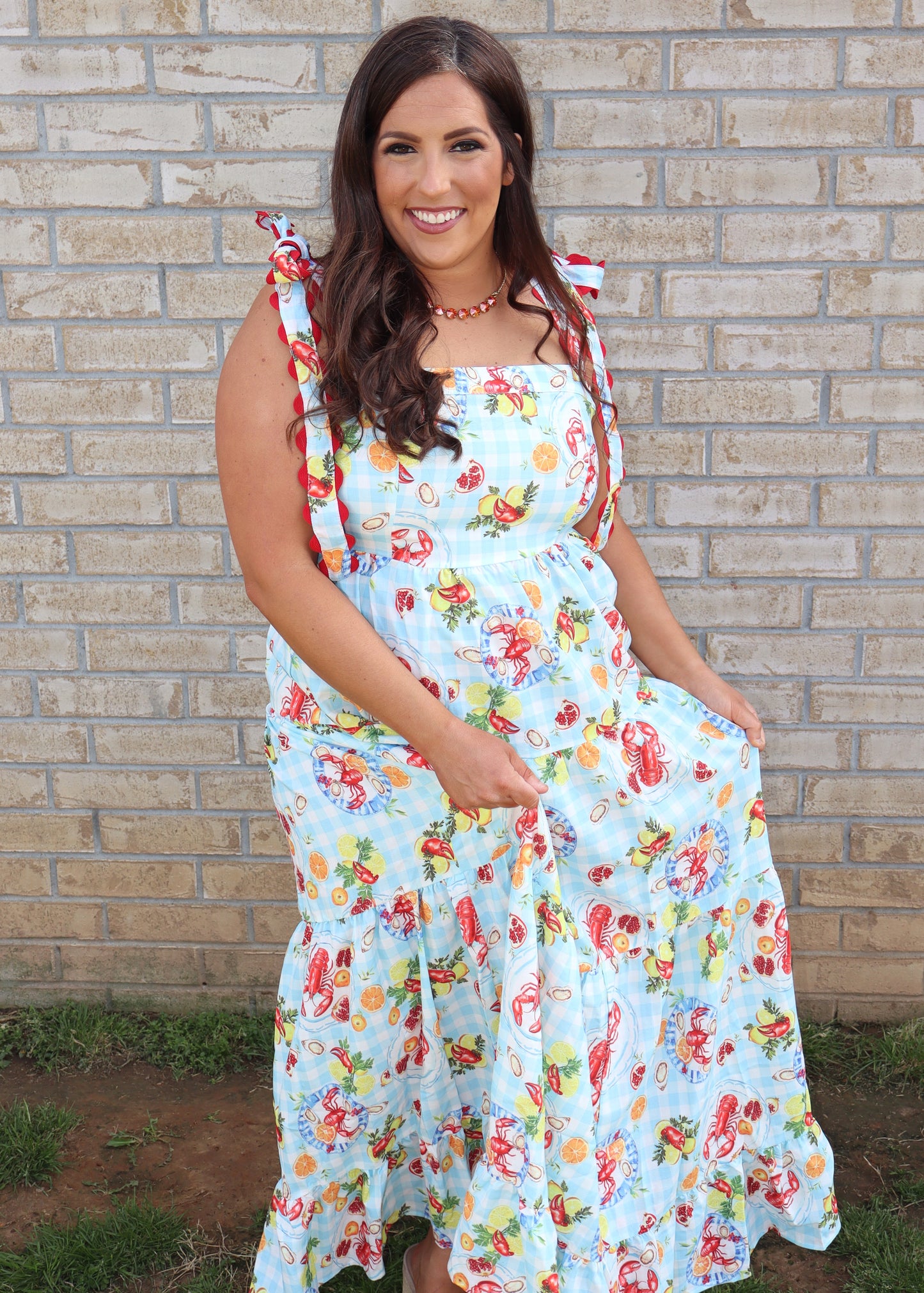 Woman wearing a blue and white gingham maxi dress with seafood boil design throughout and tie shoulder straps with bright red ric rac detail. Woman is standing in front of a brick wall.