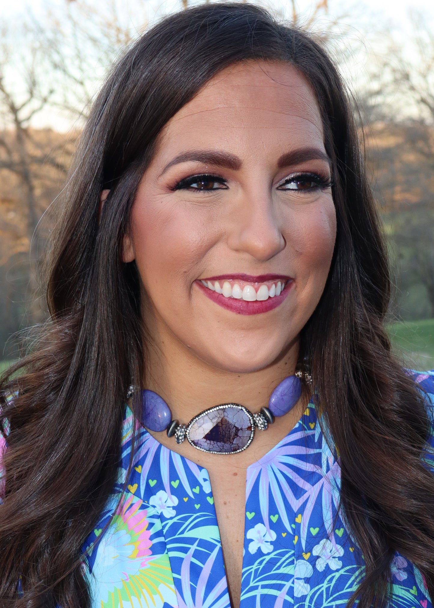 Close up of a woman wearing a tropical and parrot printed blue dress and purple choker necklace standing in a grassy field.