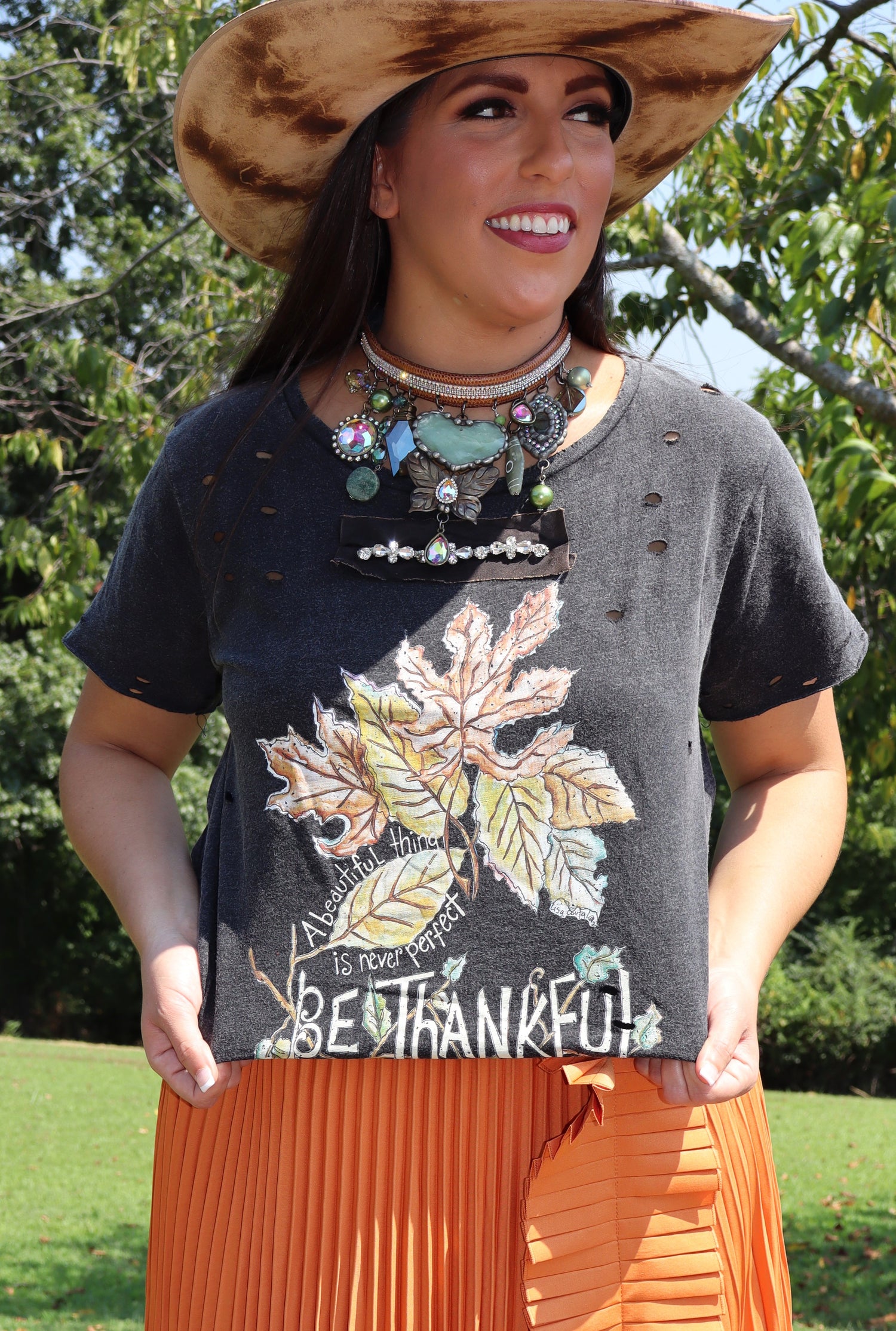 Woman wearing a graphic t-shirt with leaf design and 'Be Thankful' text, standing outdoors.