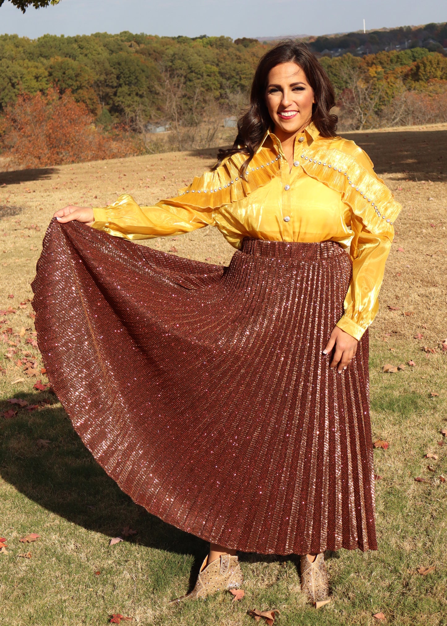 Woman wearing a yellow, shimmery blouse and bronze pleated skirt outdoors with trees in the background