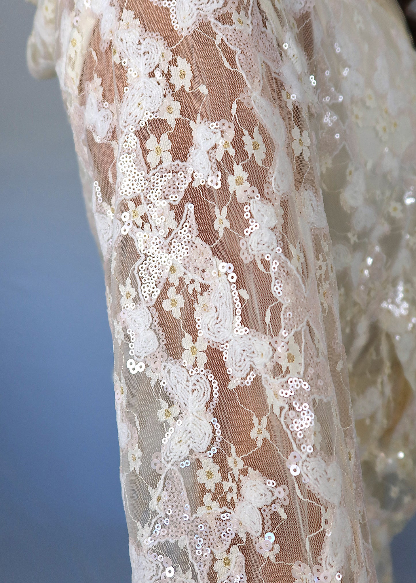 Close up of woman wearing an ivory sequin and lace, mesh jacket over a white shirt with a pink necklace against a plain background