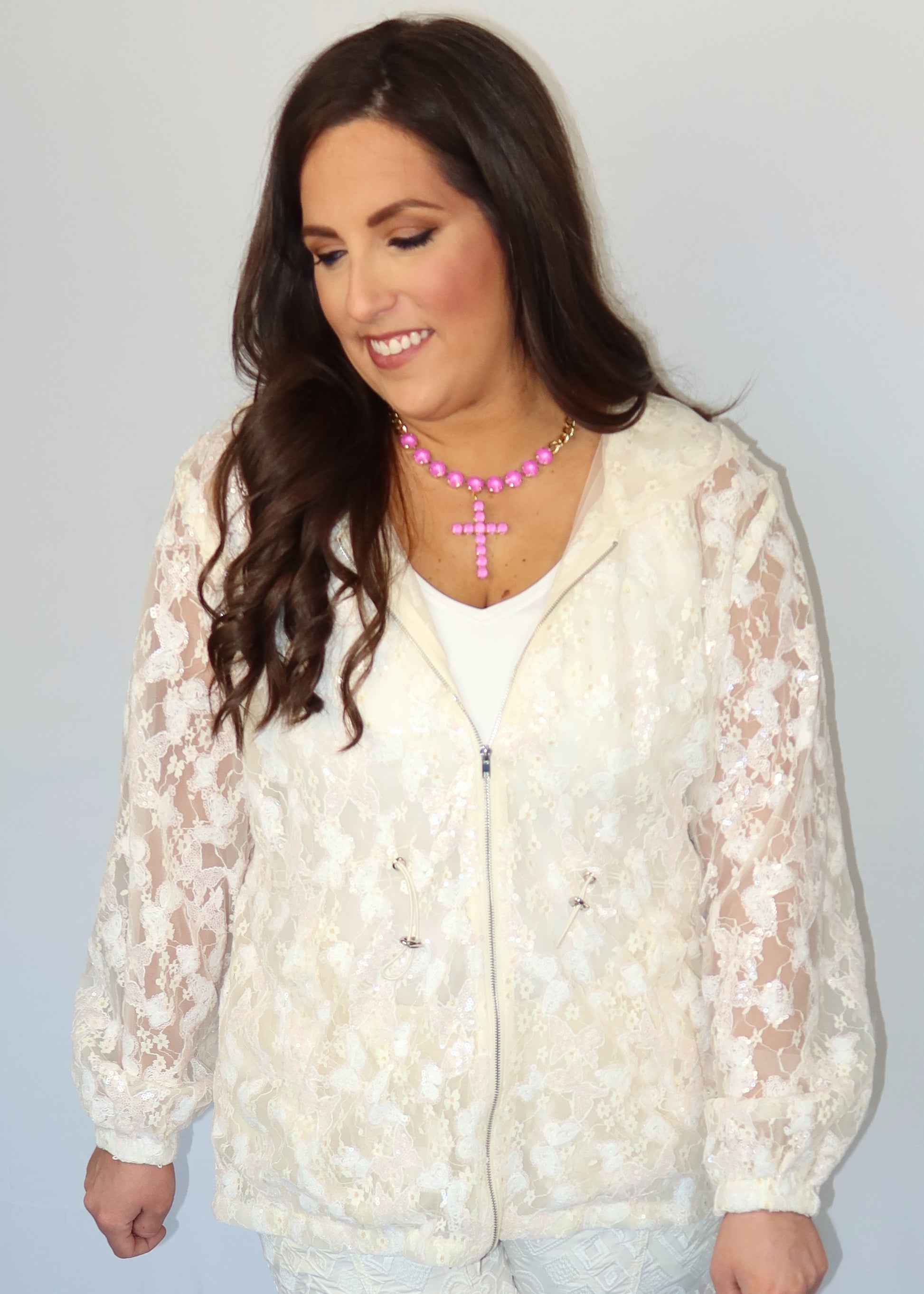 Woman wearing an ivory sequin and lace, mesh jacket over a white shirt with a pink necklace against a plain background