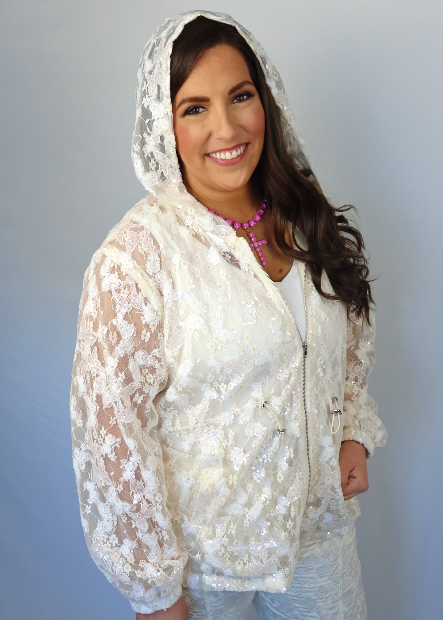 Woman wearing an ivory sequin and lace, mesh jacket over a white shirt with a pink necklace against a plain background