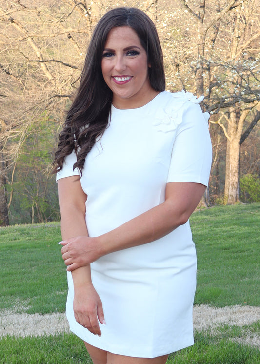 Woman wearing a white shift dress with white 3D flowers on the shoulders standing outdoors with trees and grass in the background