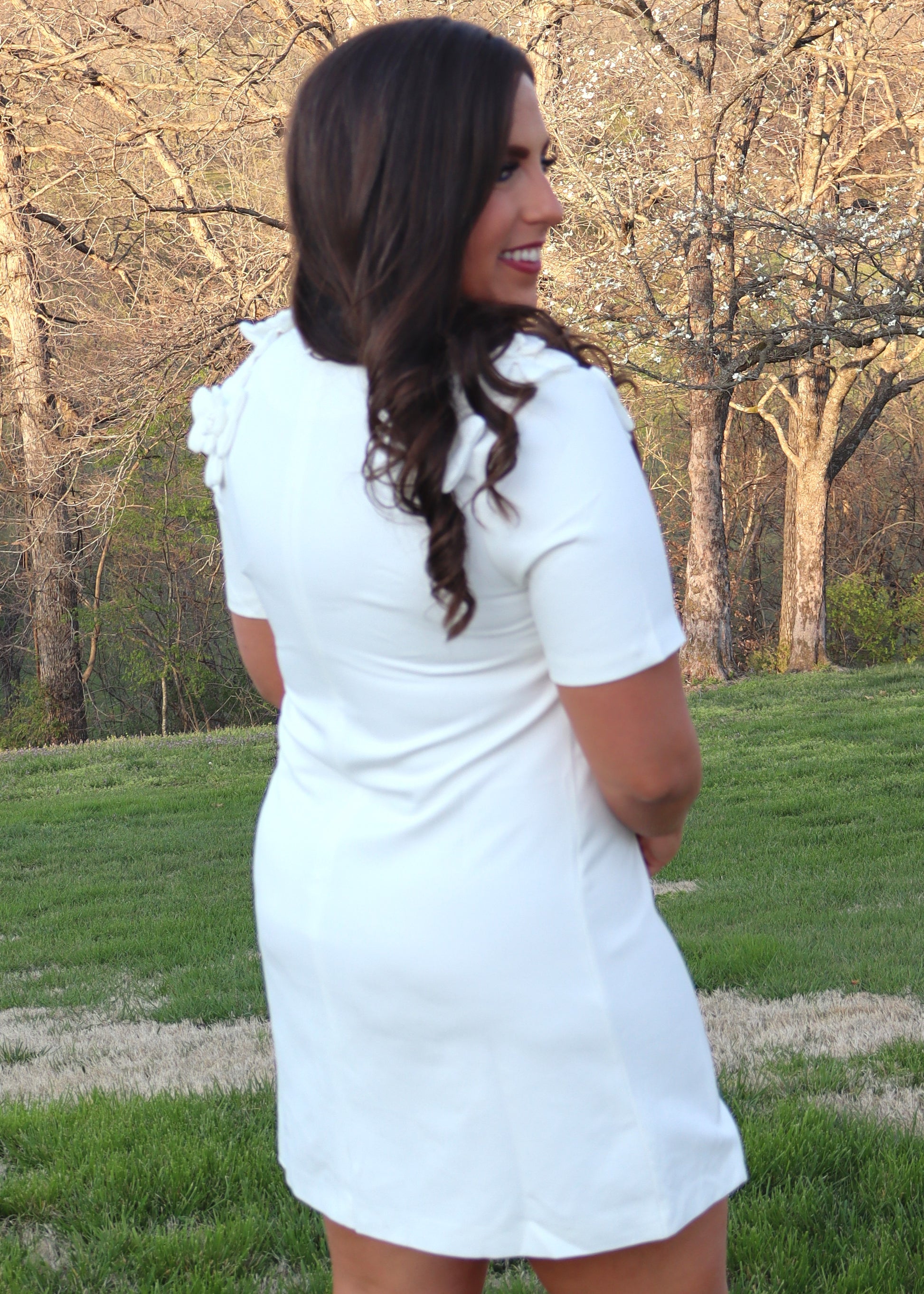 Woman wearing a white shift dress with white 3D flowers on the shoulders standing outdoors with trees and grass in the background