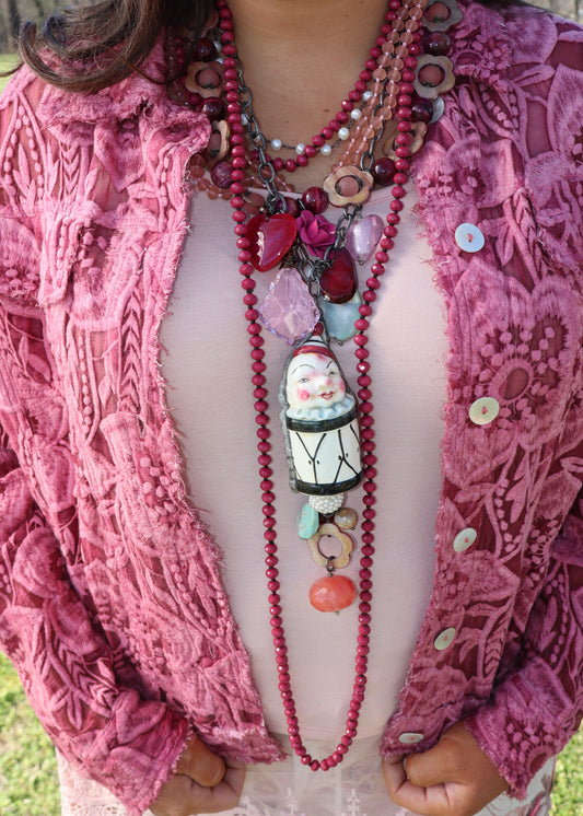 Woman wearing a long, multi-chain raspberry and various shades of pink colorful beaded necklace with a large clown drum pendant.  She is wearing a pink textured jacket. 