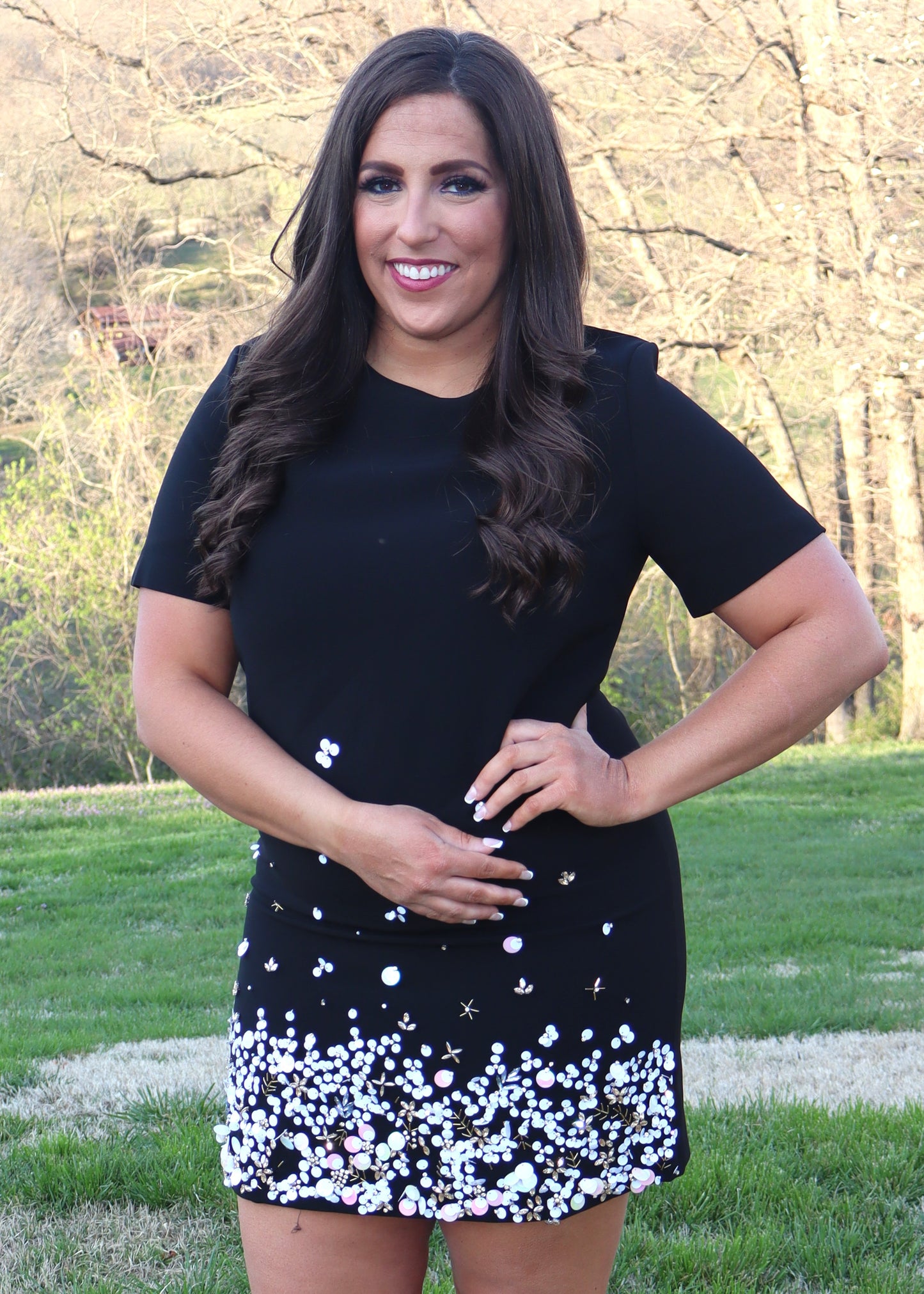 Woman wearing a black shift dress with a sequin and bead design along the bottom half of the dress and standing outside in the grass. 