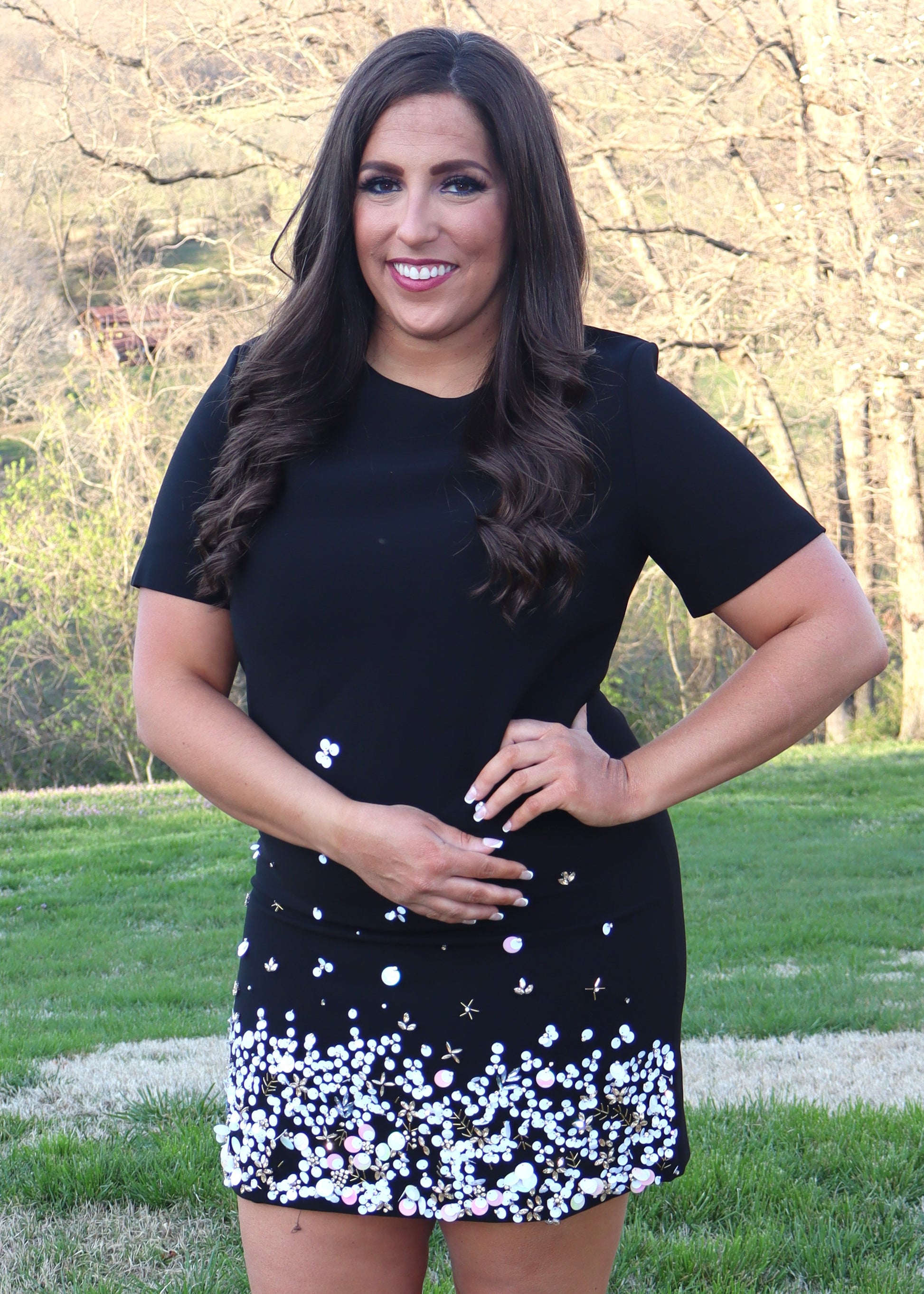 Woman wearing a black shift dress with a sequin and bead design along the bottom half of the dress and standing outside in the grass. 