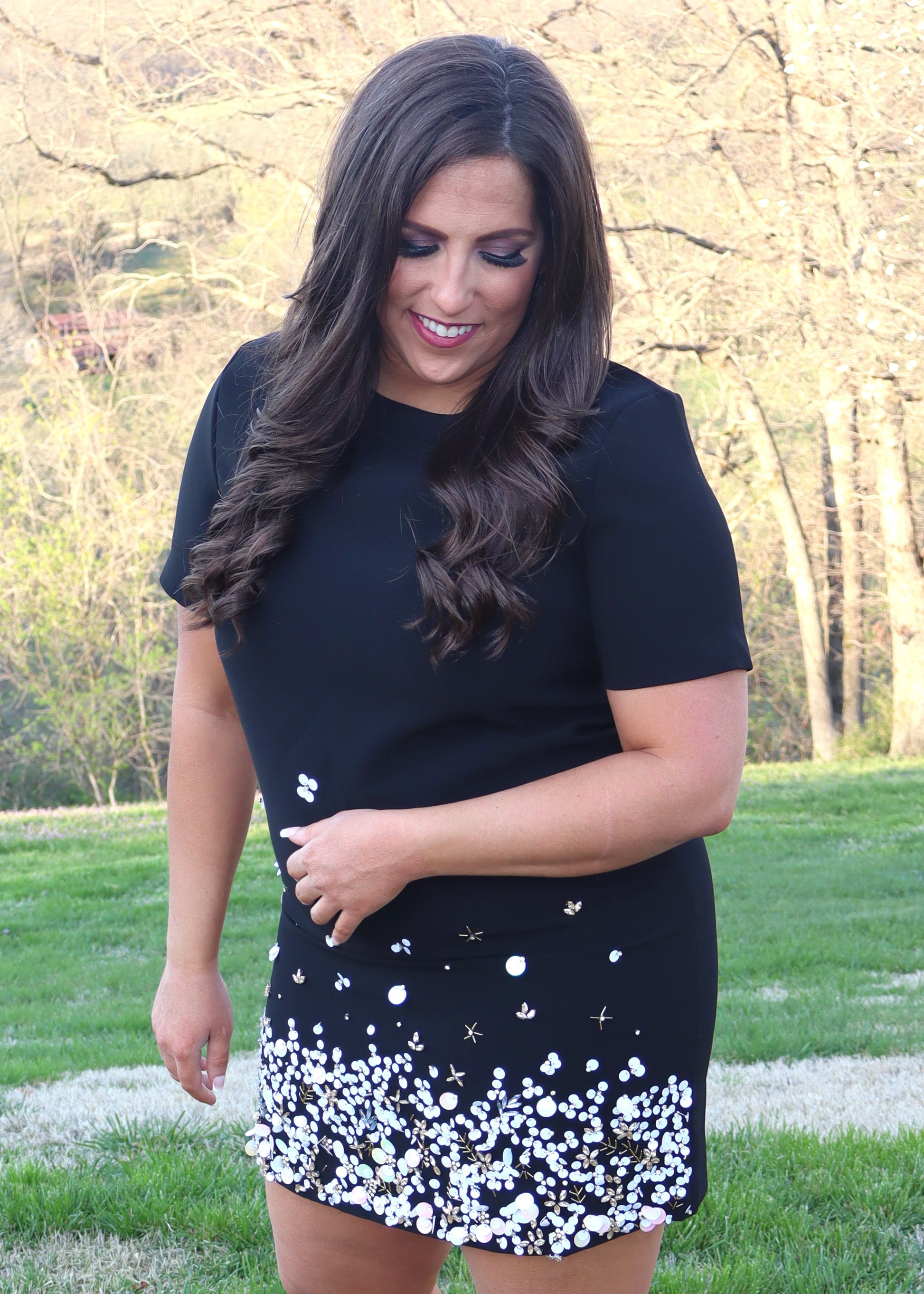 Woman wearing a black shift dress with a sequin and bead design along the bottom half of the dress and standing outside in the grass. 