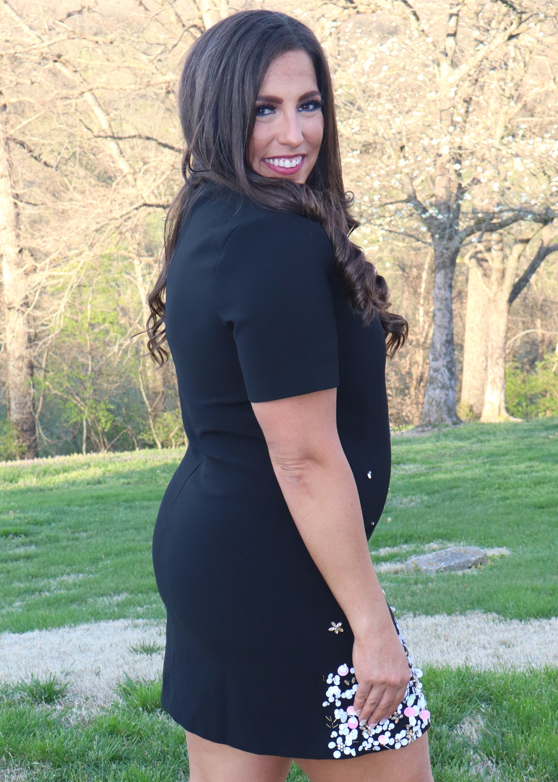 Woman wearing a black shift dress with a sequin and bead design along the bottom half of the dress and standing outside in the grass. 
