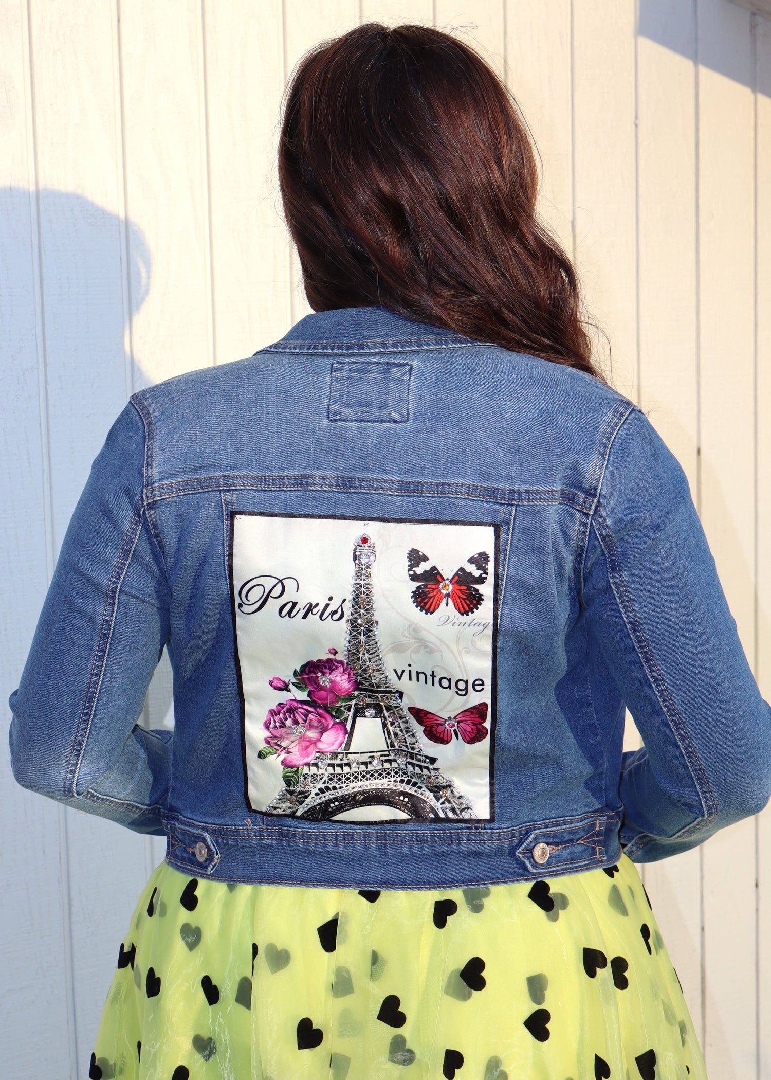 Woman wearing a denim jacket with a graphic design of the Eiffel Tower and butterflies on the back and 'Paris' and 'vintage' text, standing against a light wooden panel background.