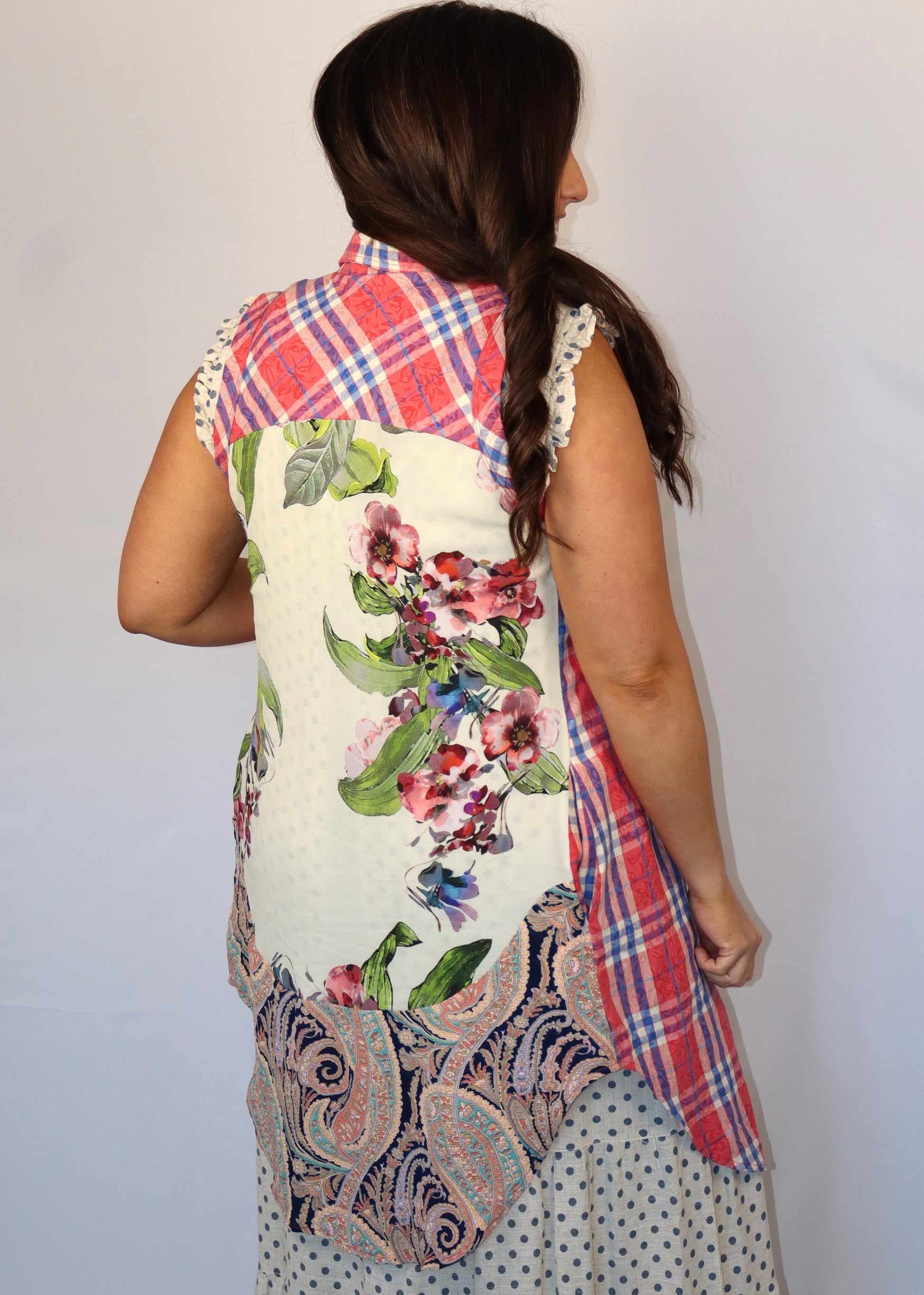 Woman wearing a colorful, patterned tunic top with a floral and plaid design over a polka dot dress while next to a plain background
