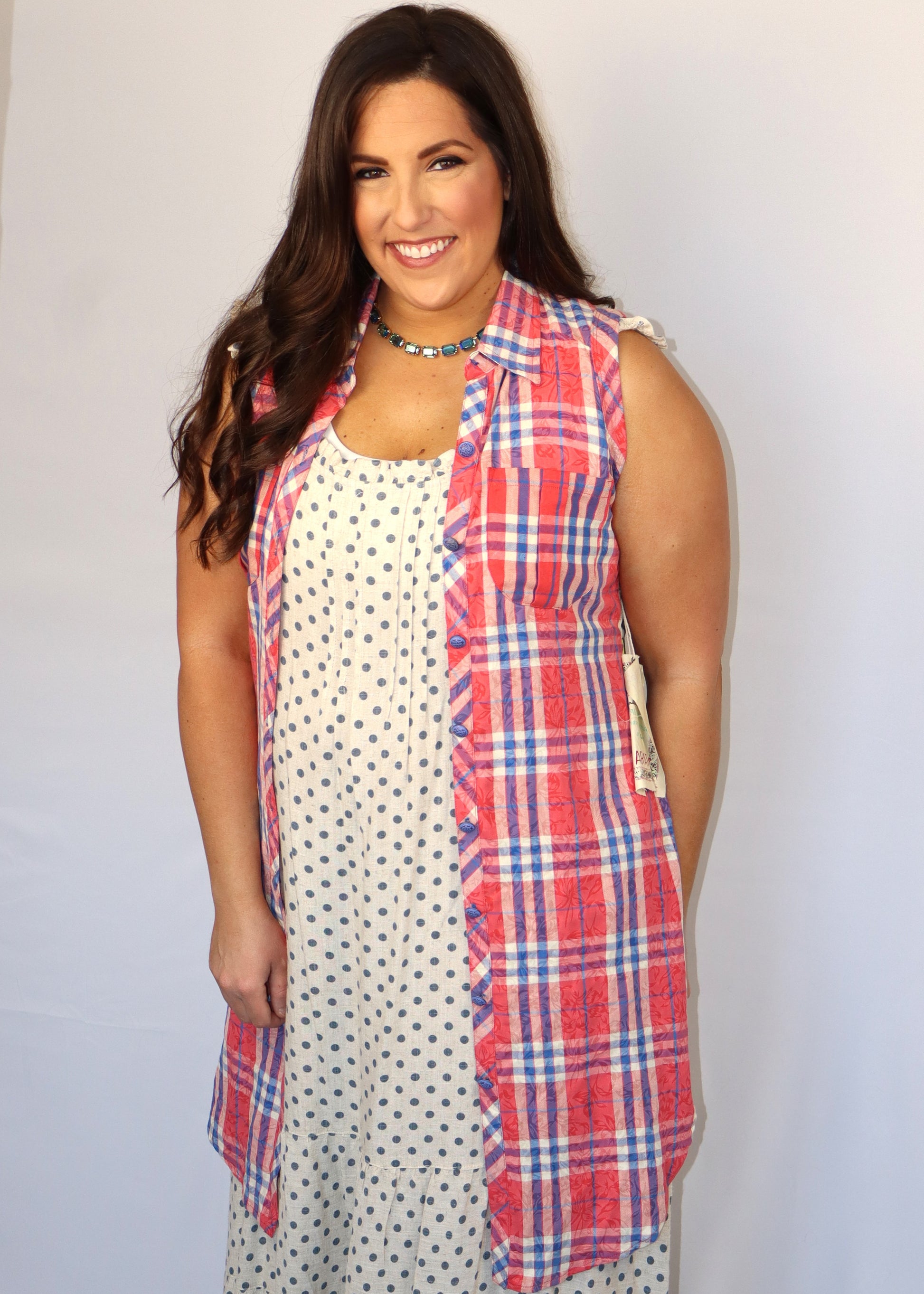 Woman wearing a red and blue plaid tunic top over a polka dot dress while next to a plain background