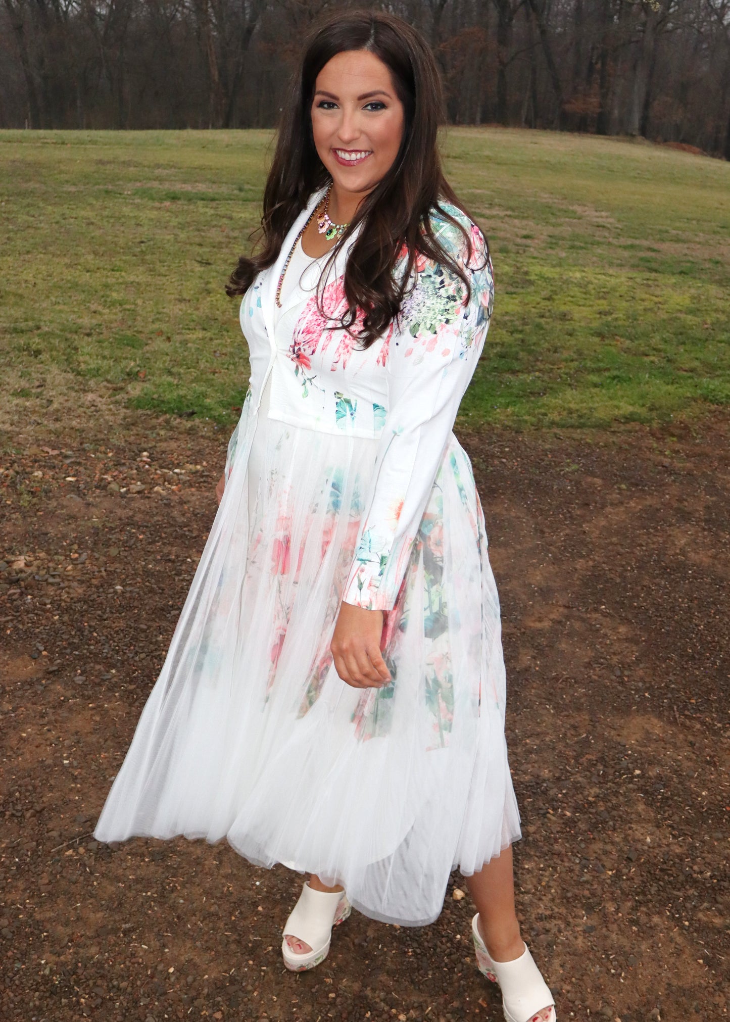 Woman wearing a white floral duster standing in a natural setting with grass and dirt.