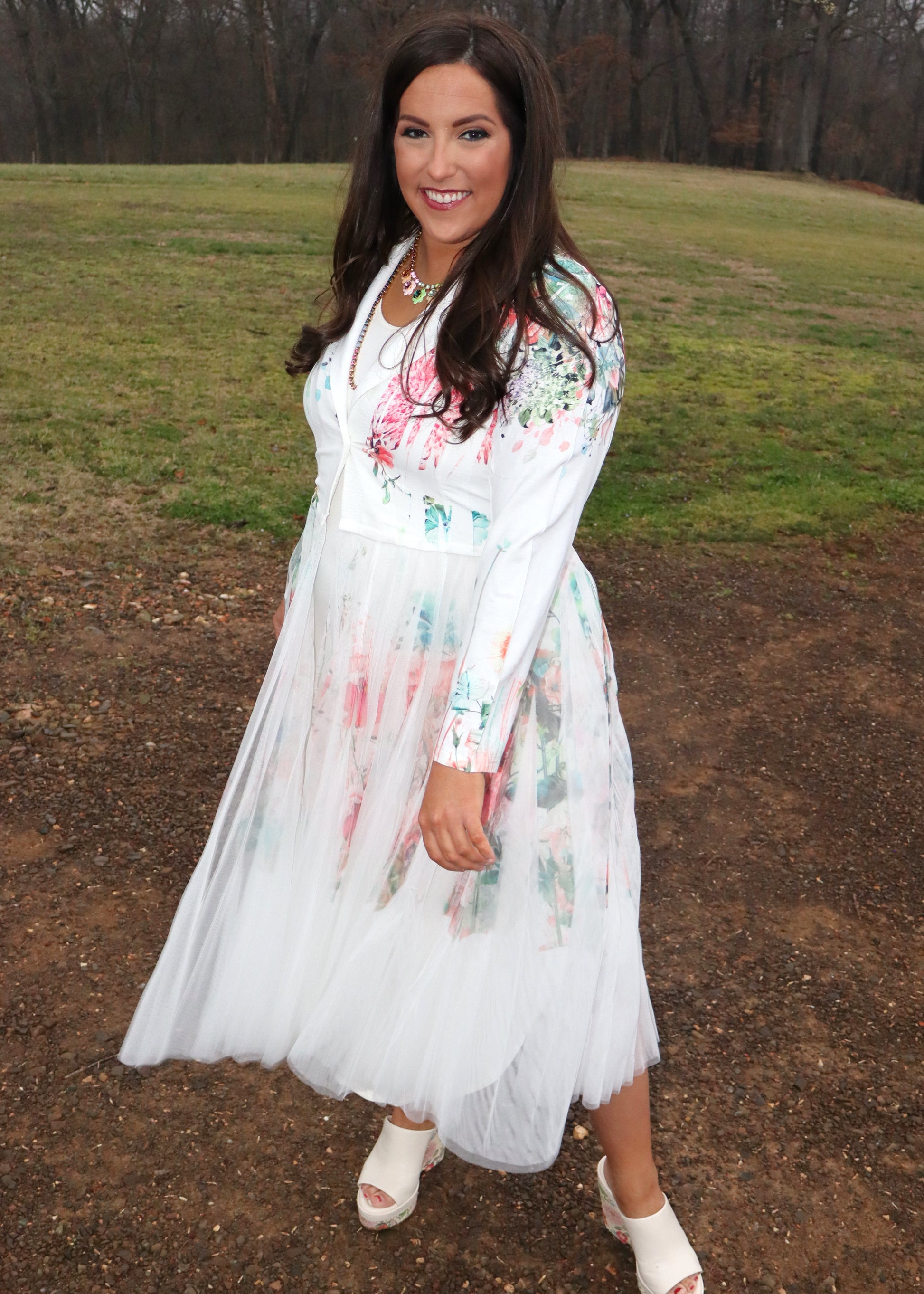 Woman wearing a white floral duster standing in a natural setting with grass and dirt.