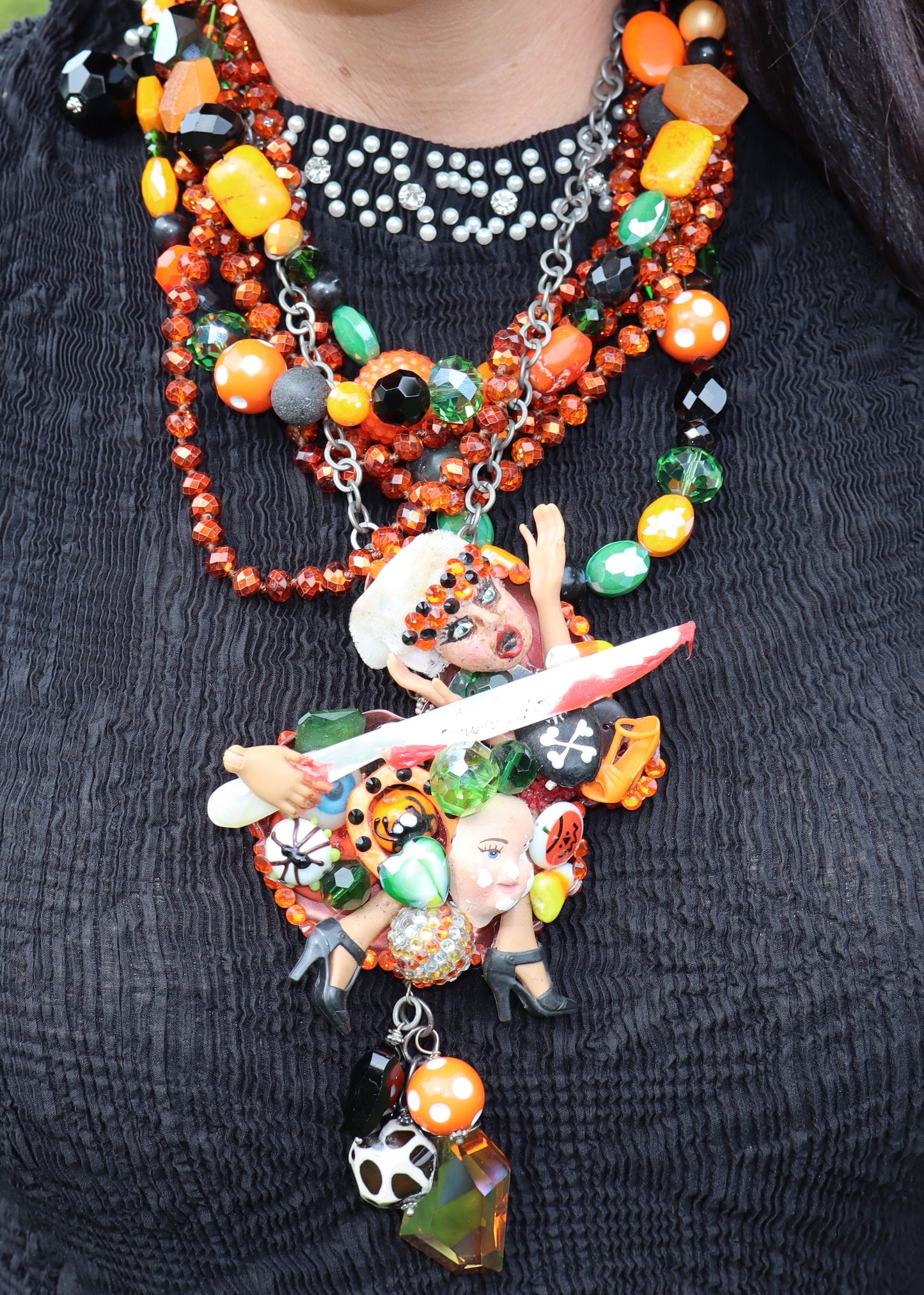 Orange, green, and black multi-strand beaded Art by Amy upcycled necklace with Halloween, scary movie-themed charms on a black stand against a grassy background