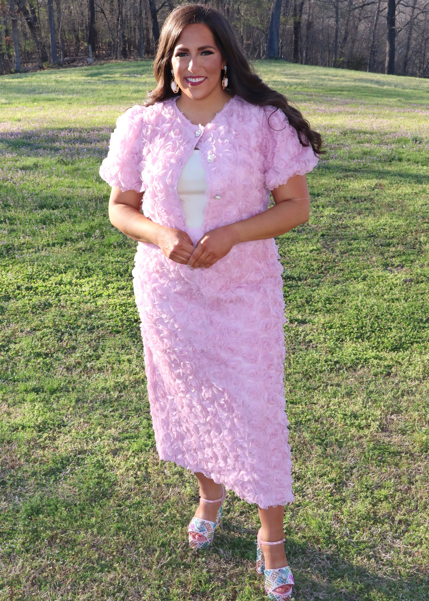Woman wearing a pink matching skirt and tops with 3D floral sleeves standing in a grassy field.