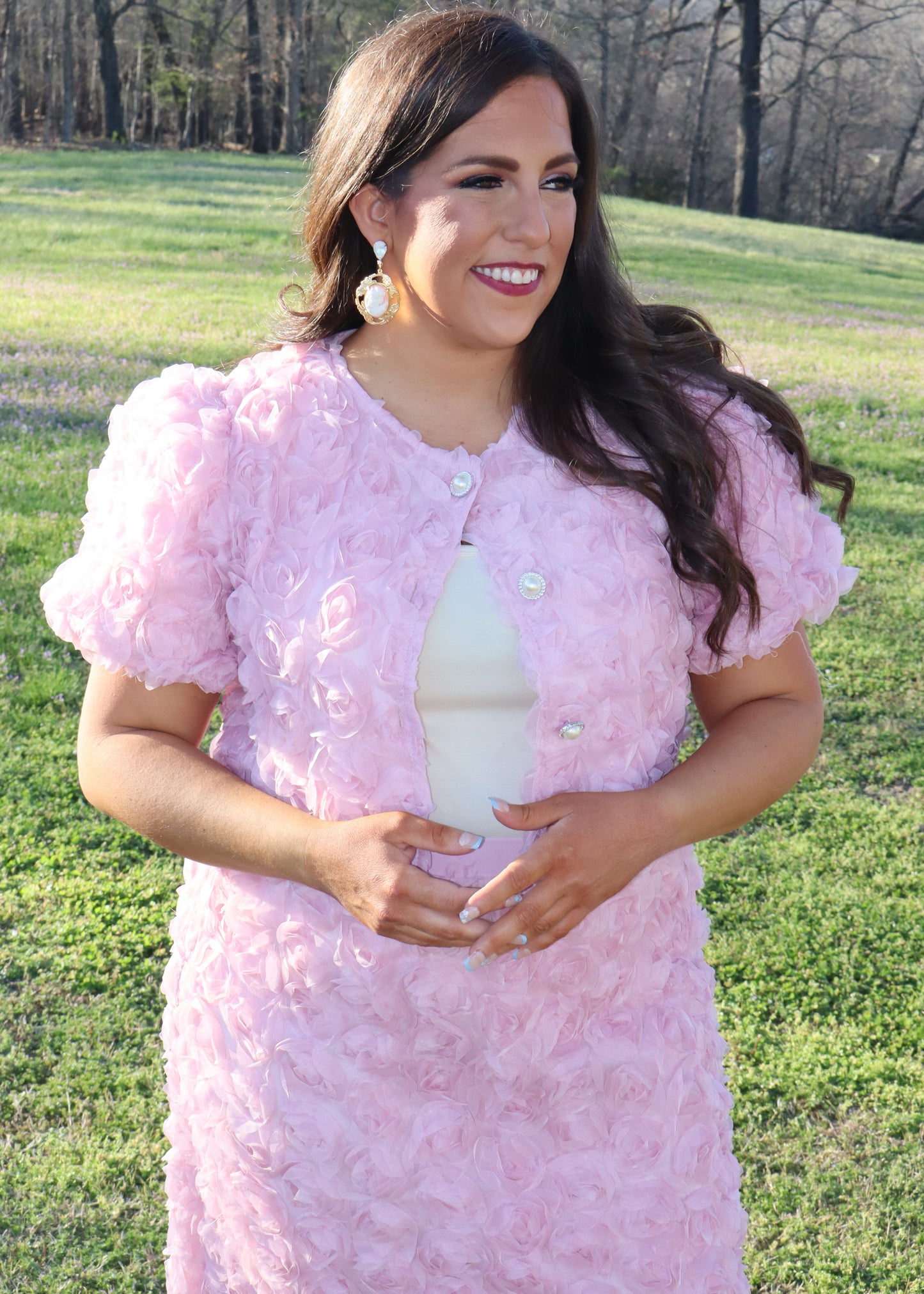 Woman wearing a pink matching skirt and tops with 3D floral sleeves standing in a grassy field.