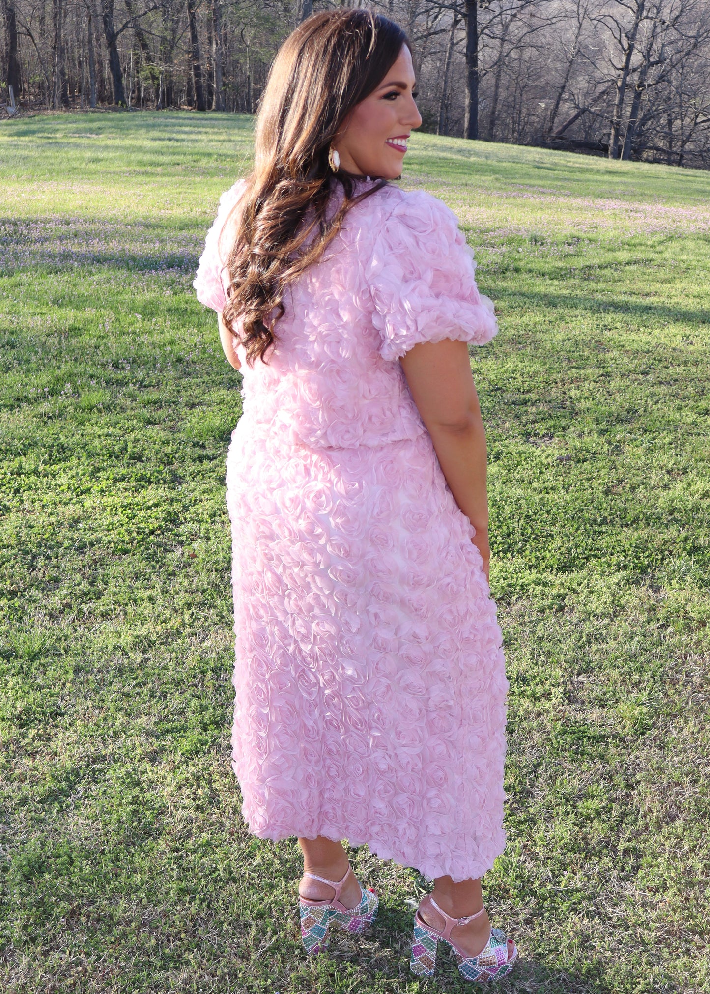 Woman wearing a pink matching skirt and tops with 3D floral sleeves standing in a grassy field.