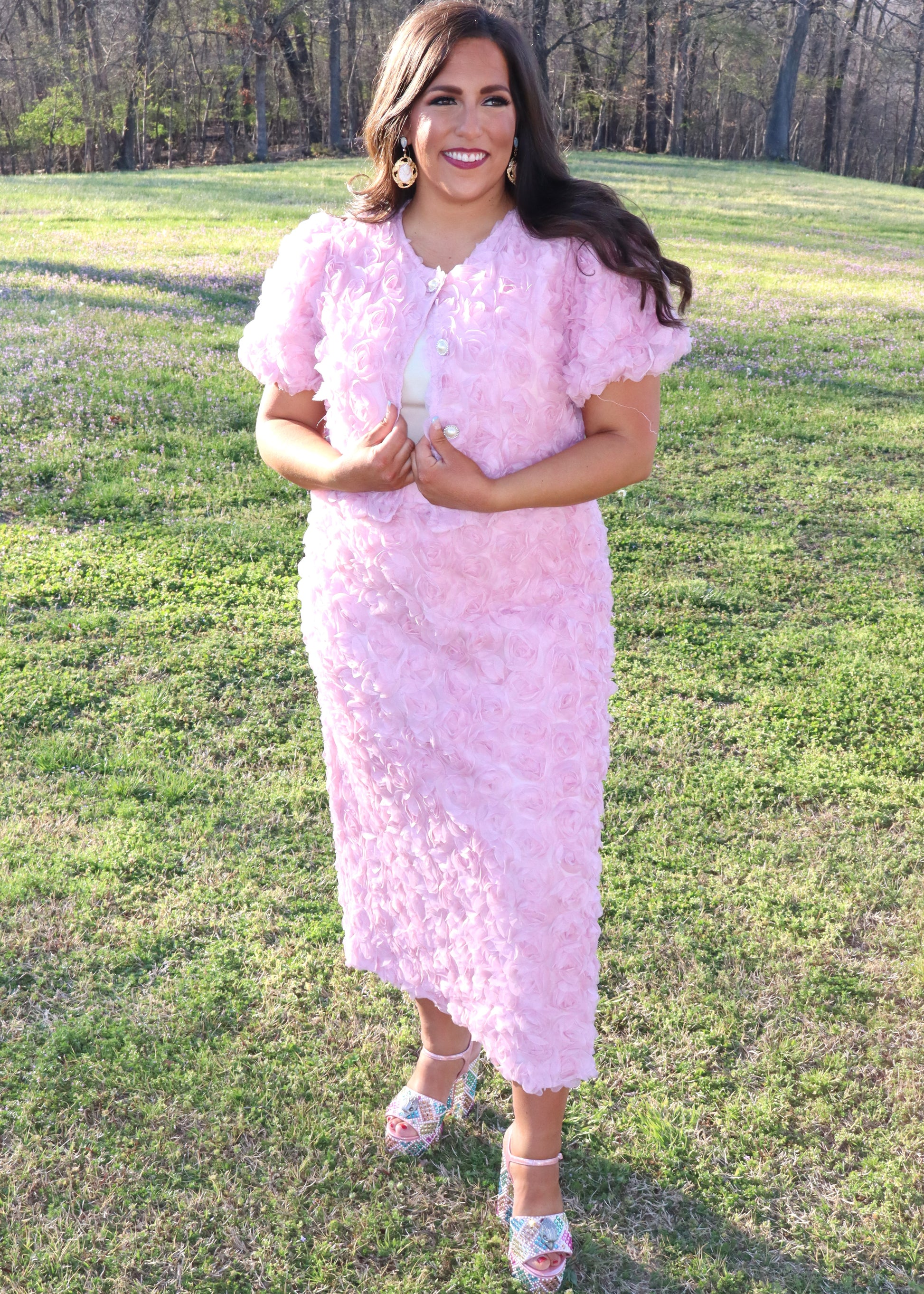Woman wearing a pink matching skirt and tops with 3D floral sleeves standing in a grassy field.