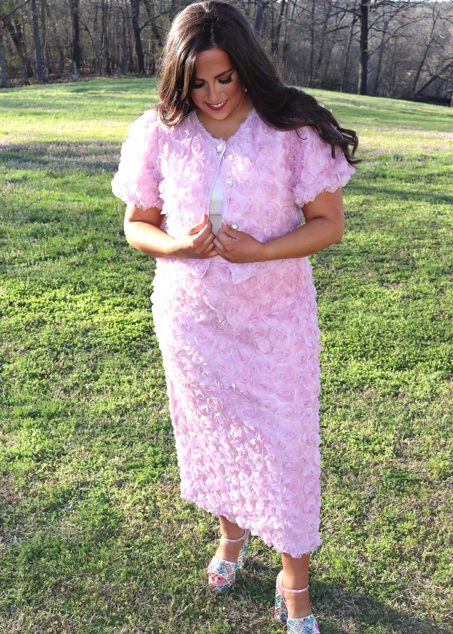 Woman wearing a pink matching skirt and tops with 3D floral sleeves standing in a grassy field.