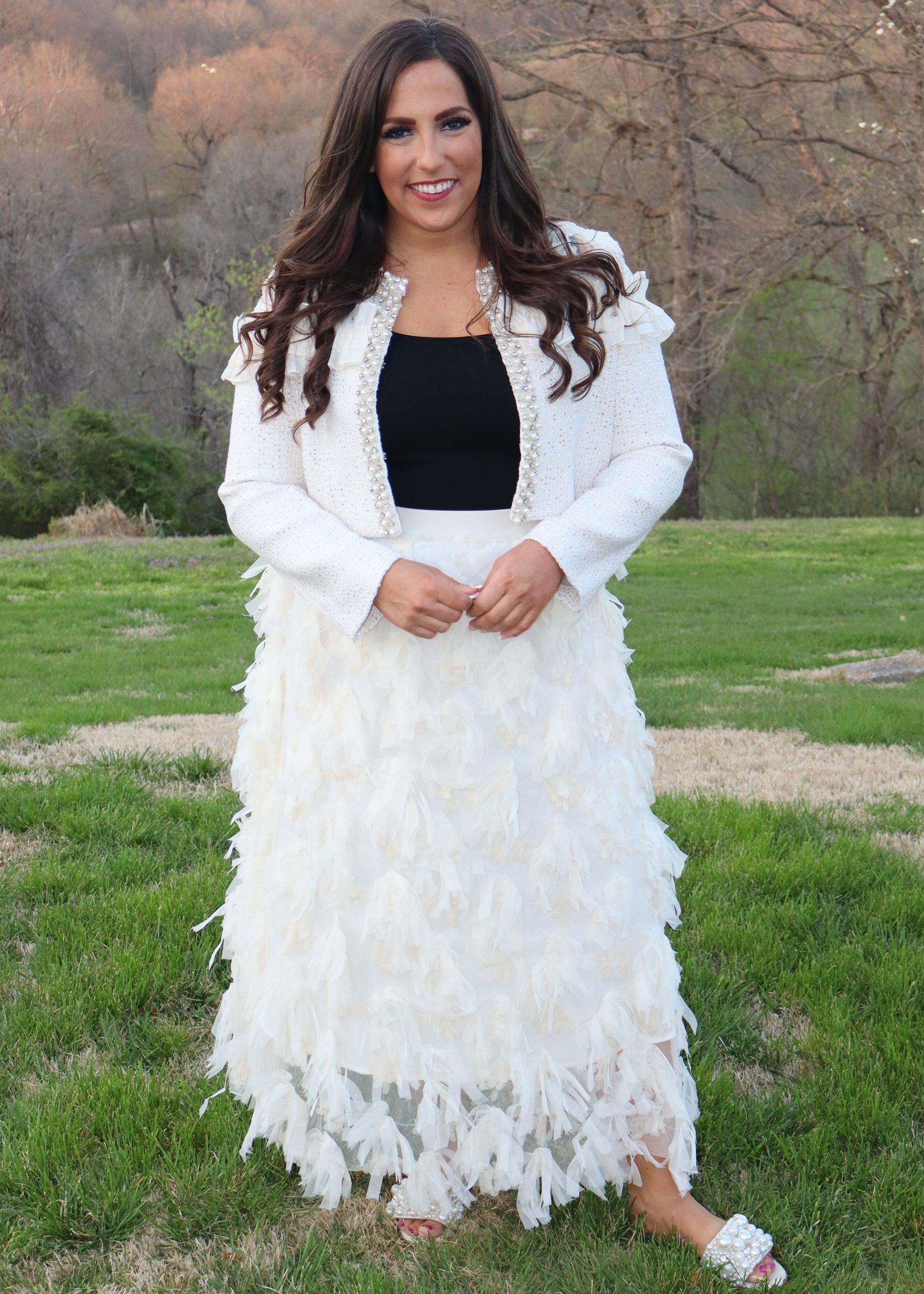 Woman wearing an ivory feathered skirt, ivory jacket, and with a black top outdoors.