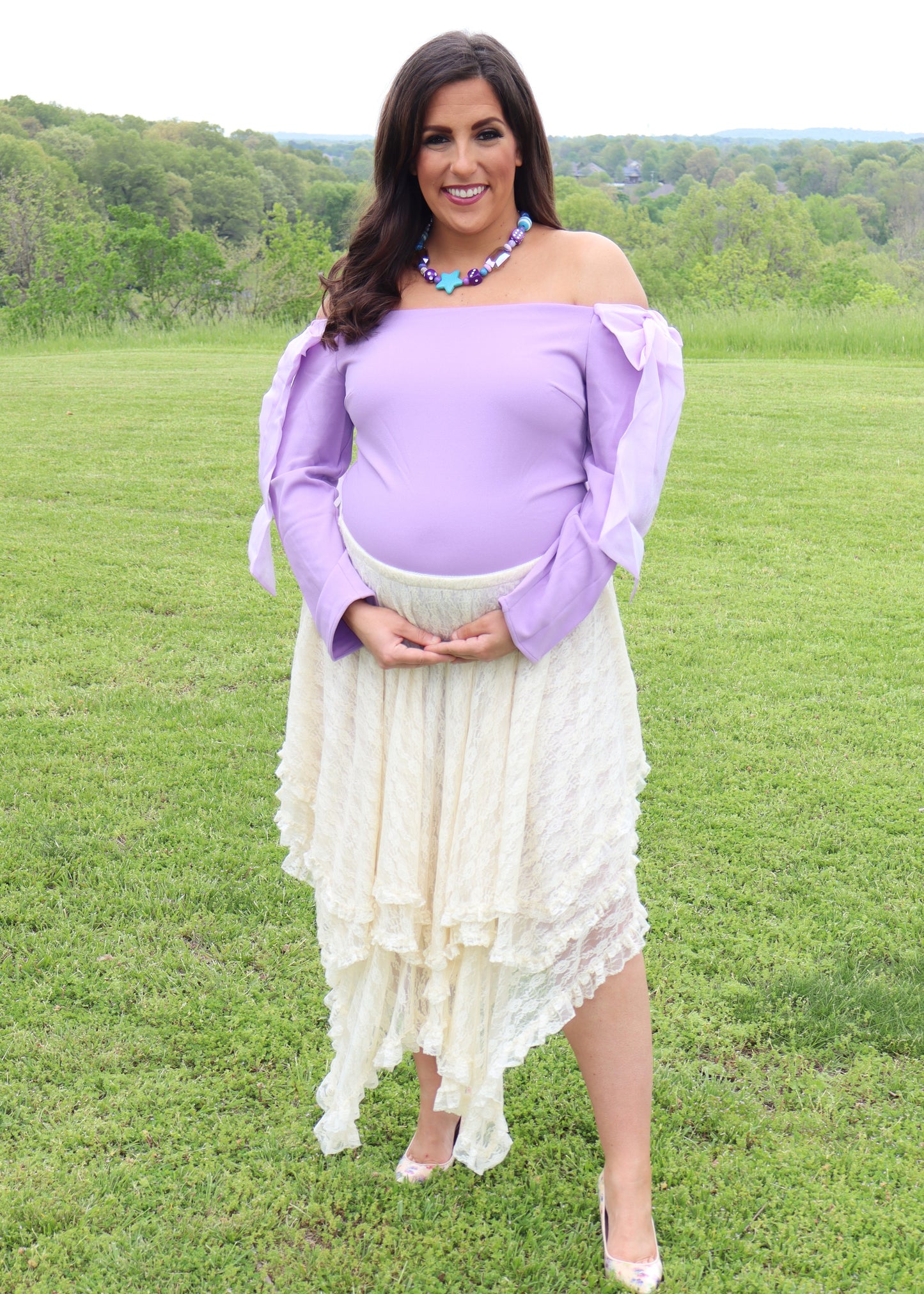 Woman in an off-the-shoulder light purple top and cream lace skirt standing in a grassy field.