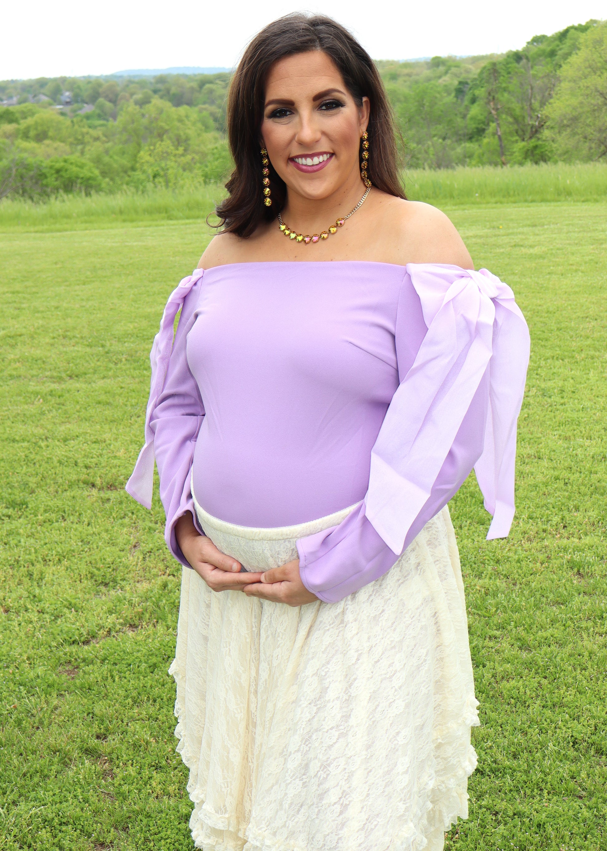 Woman in an off-the-shoulder light purple top and cream lace skirt standing in a grassy field.