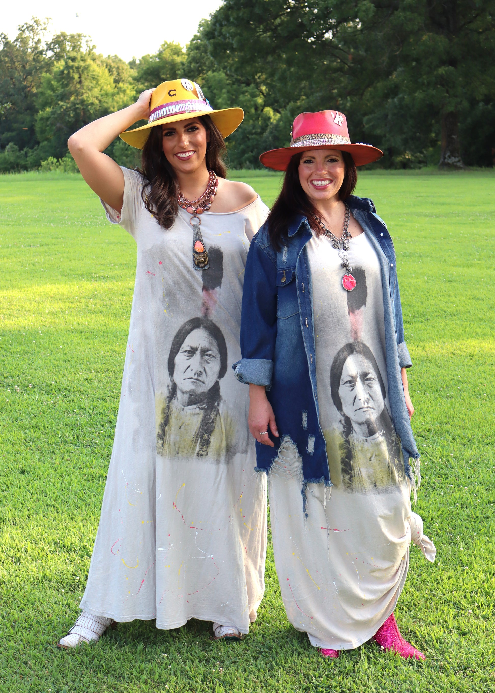 Two women wearing wide-neck dresses with an American Indian face design on a grassy field.
