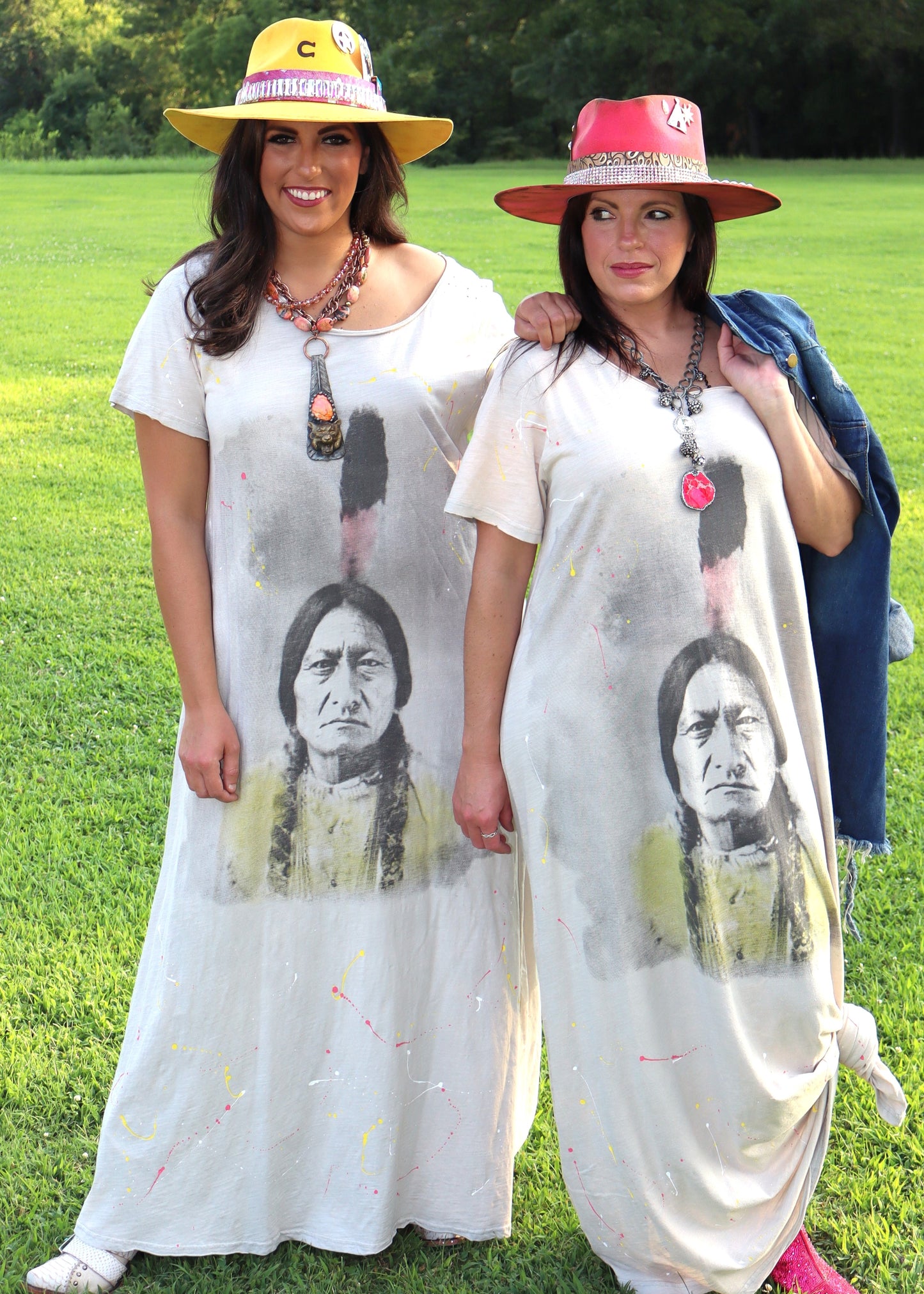 Two women wearing wide-neck dresses with an American Indian face design on a grassy field.