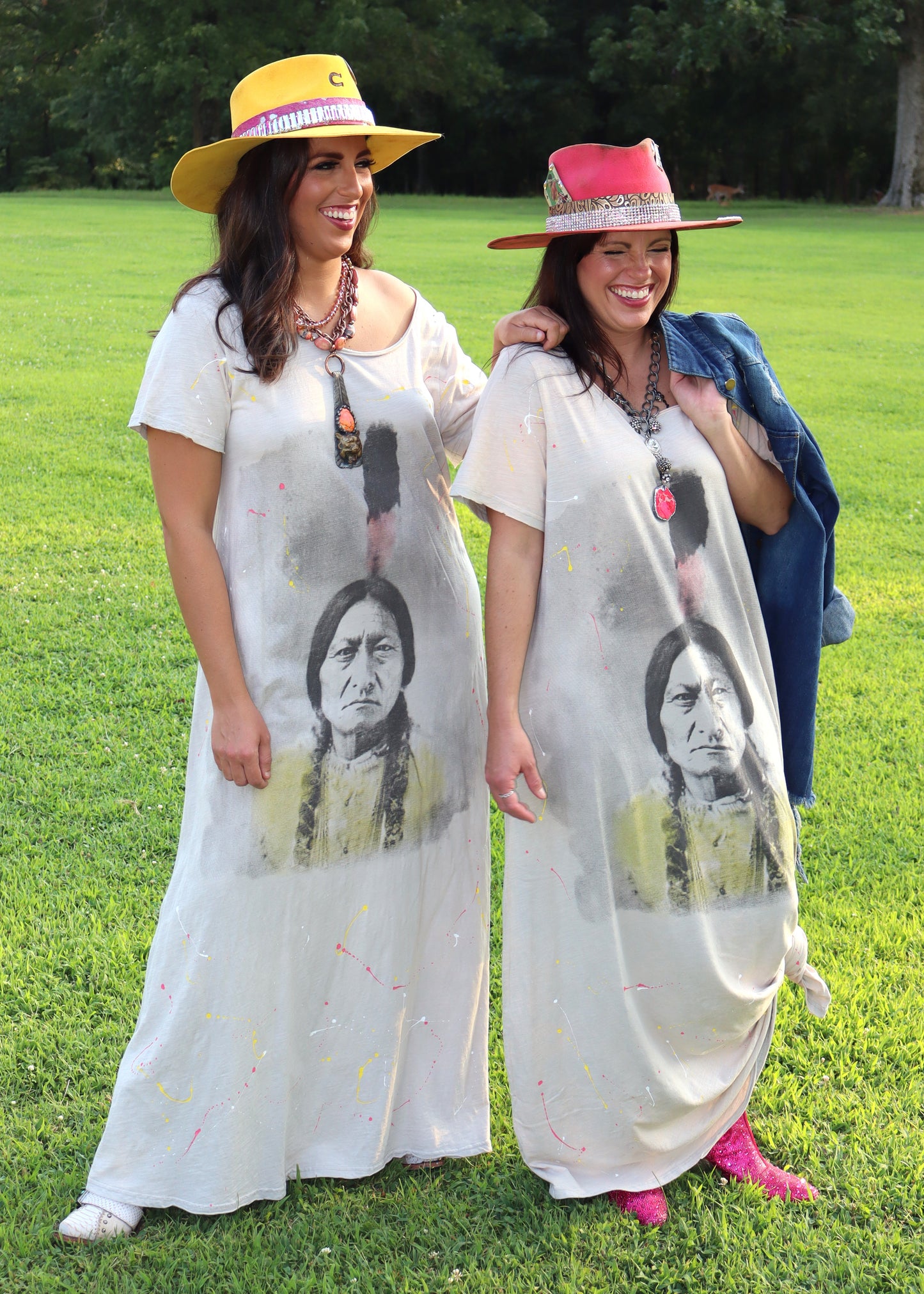 Two women wearing wide-neck dresses with an American Indian face design on a grassy field.