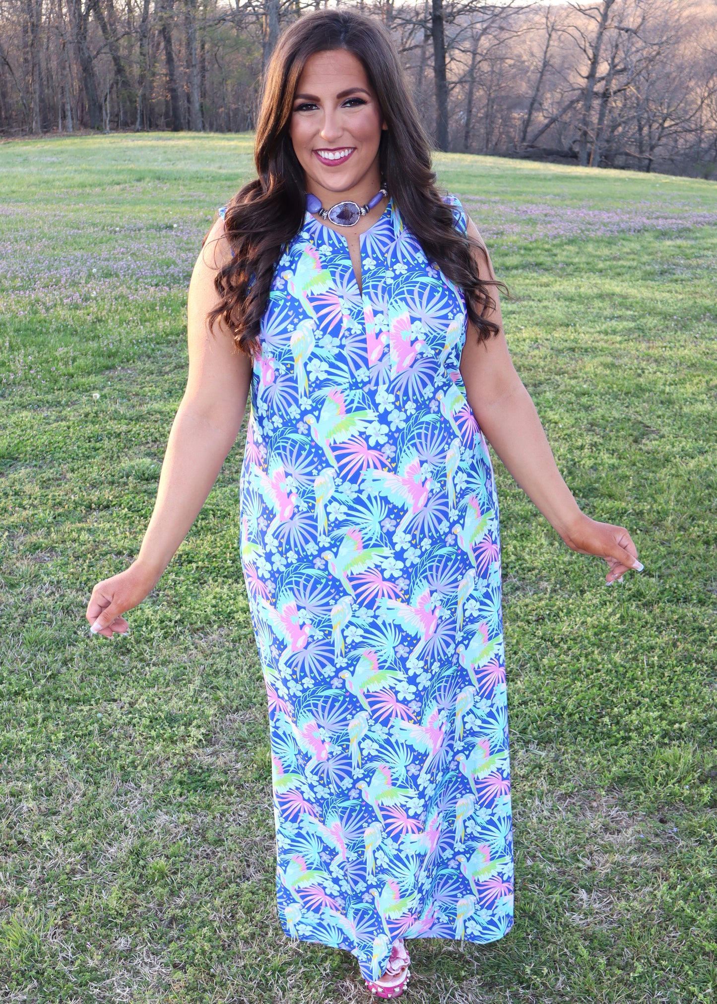 Woman wearing a tropical and parrot printed blue dress standing in a grassy field.