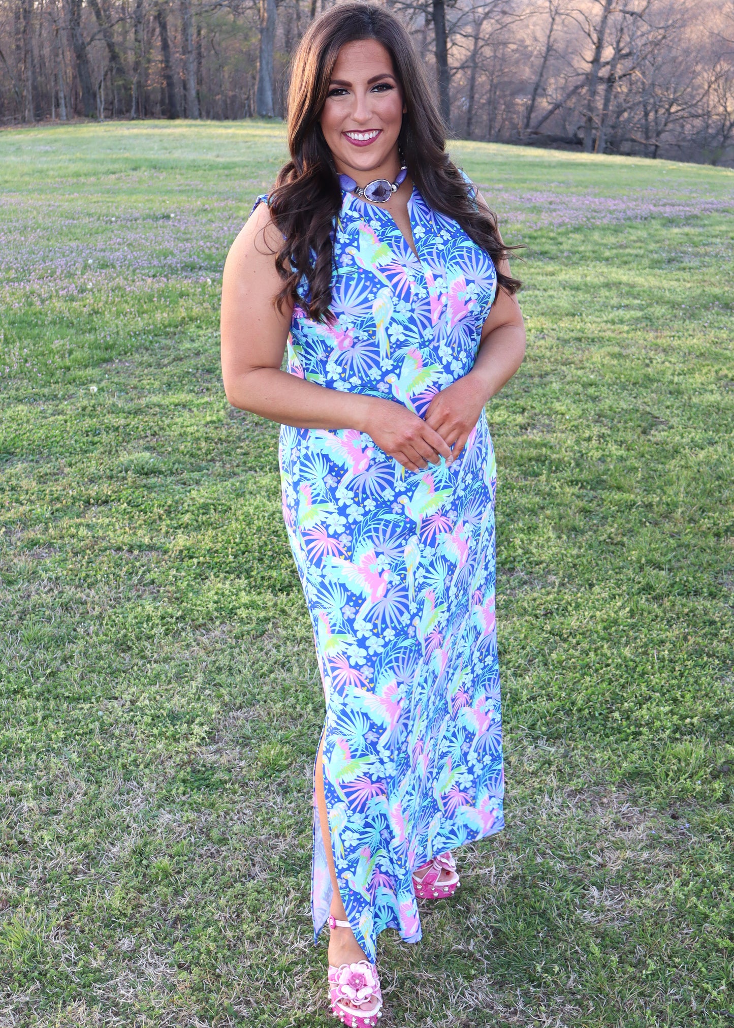 Woman wearing a tropical and parrot printed blue dress standing in a grassy field.