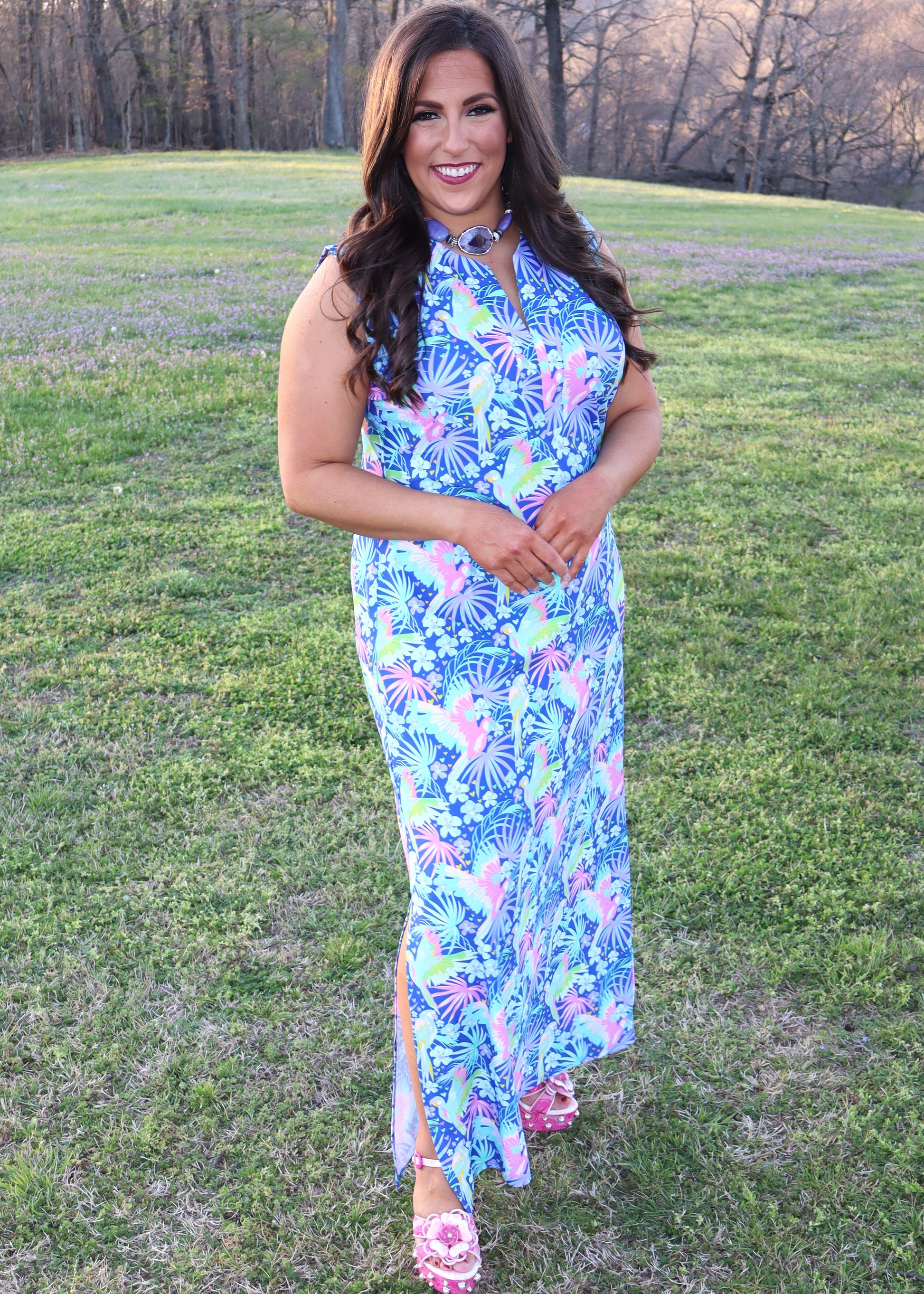 Woman wearing a tropical and parrot printed blue dress standing in a grassy field.