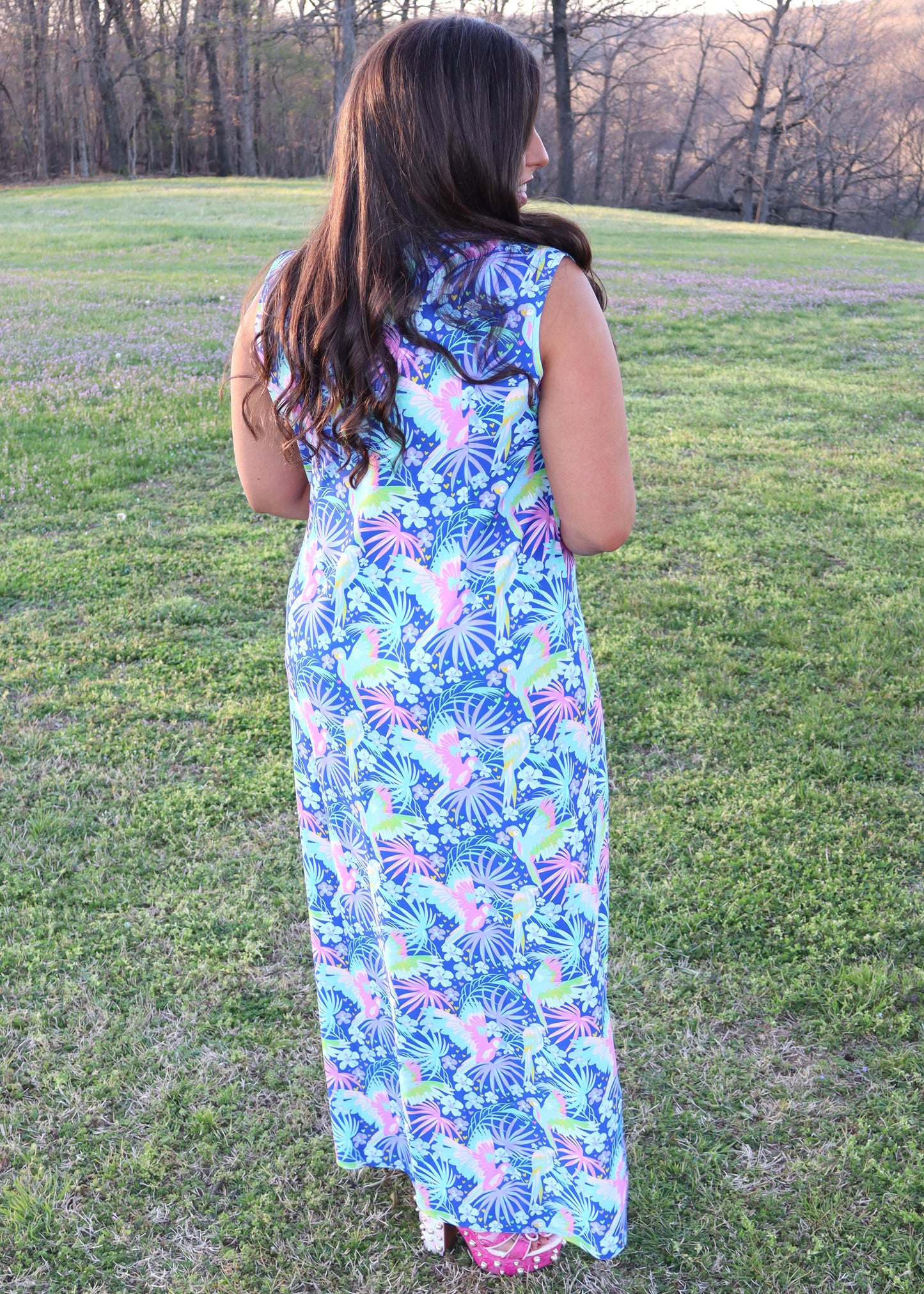 Woman wearing a tropical and parrot printed blue dress standing in a grassy field.