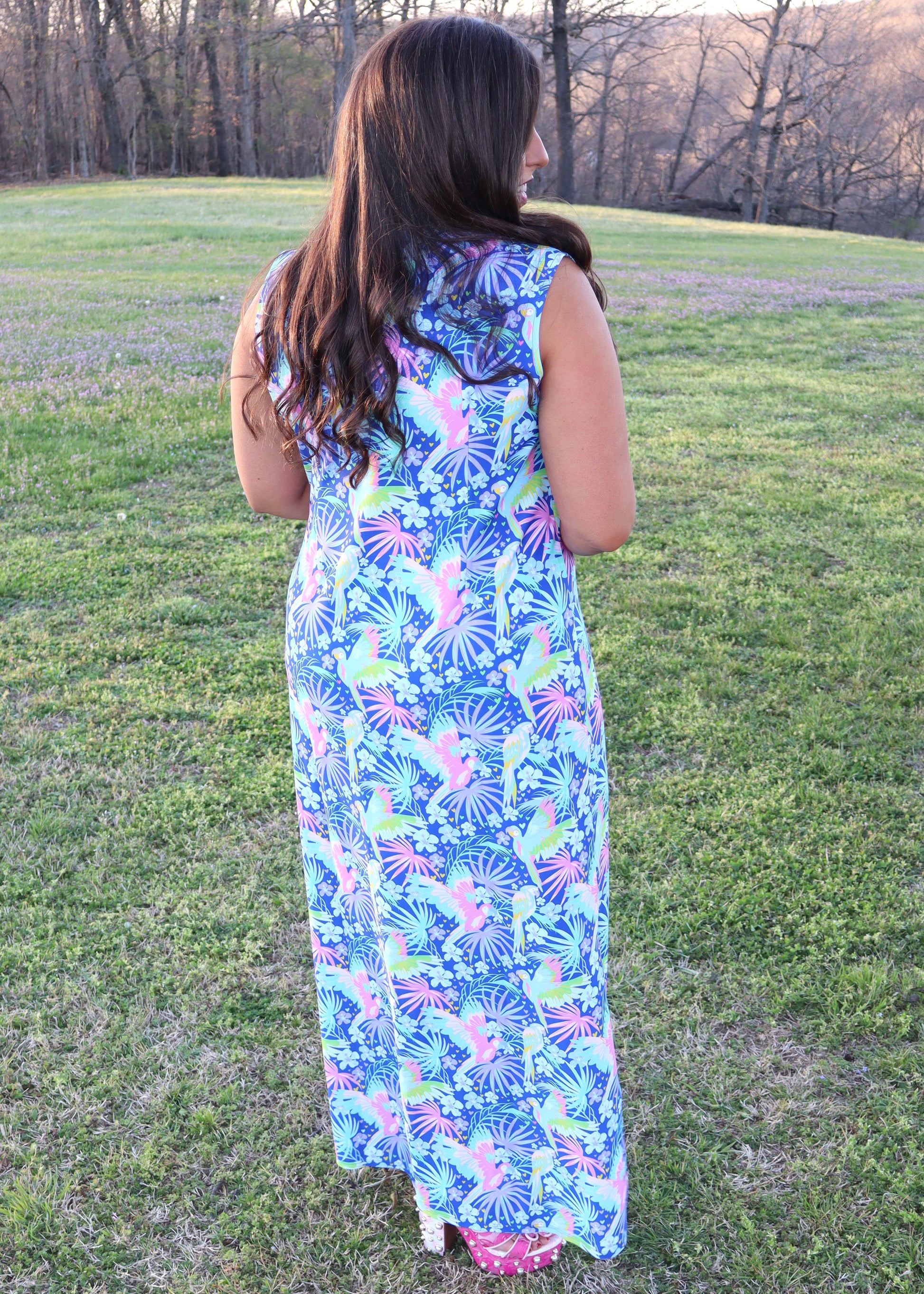 Woman wearing a tropical and parrot printed blue dress standing in a grassy field.