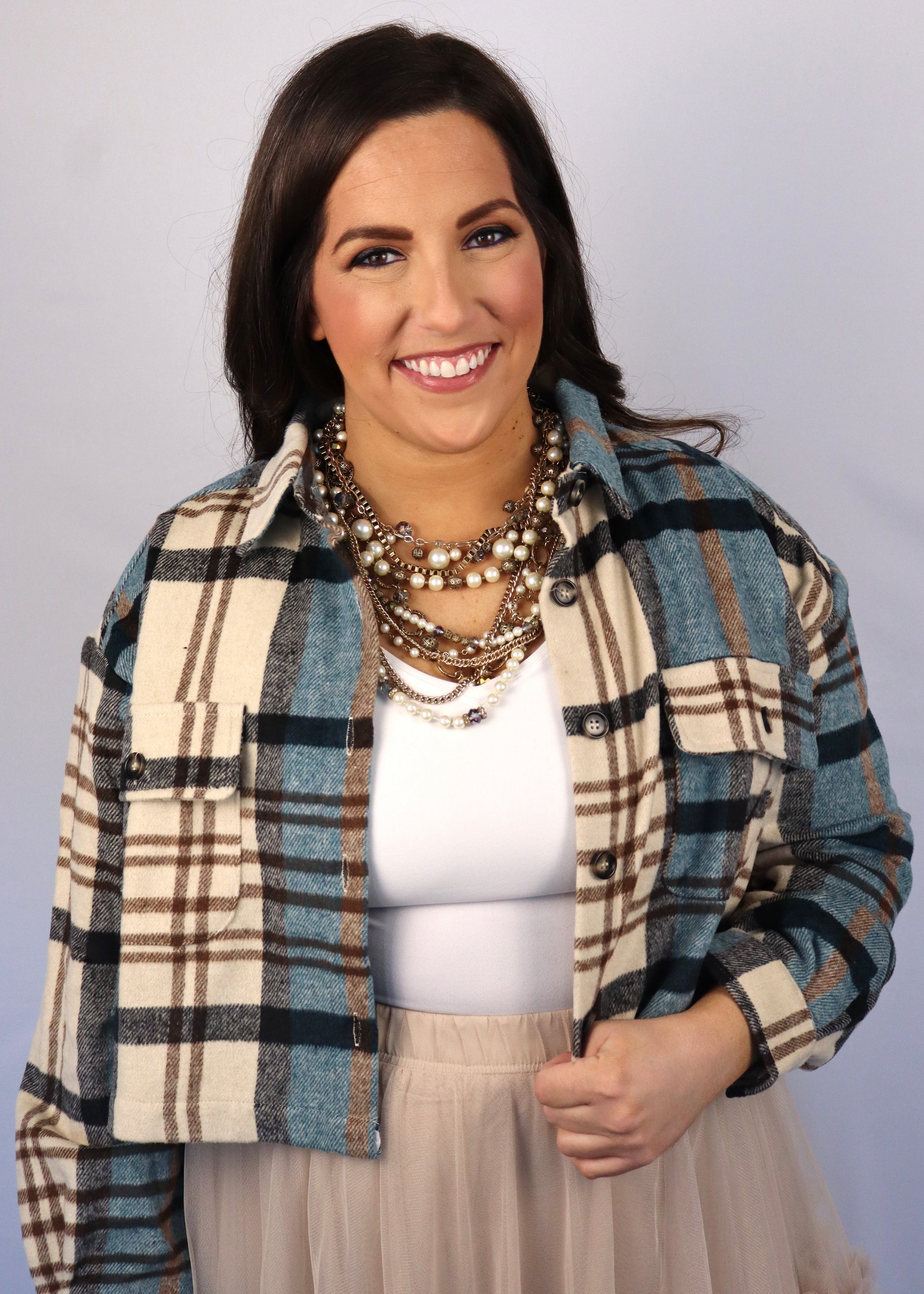 Woman wearing a teal, brown, and black cropped flannel jacket with a white tank top, taupe tiered tulle skirt, and multi-strand pearl necklace against a plain background