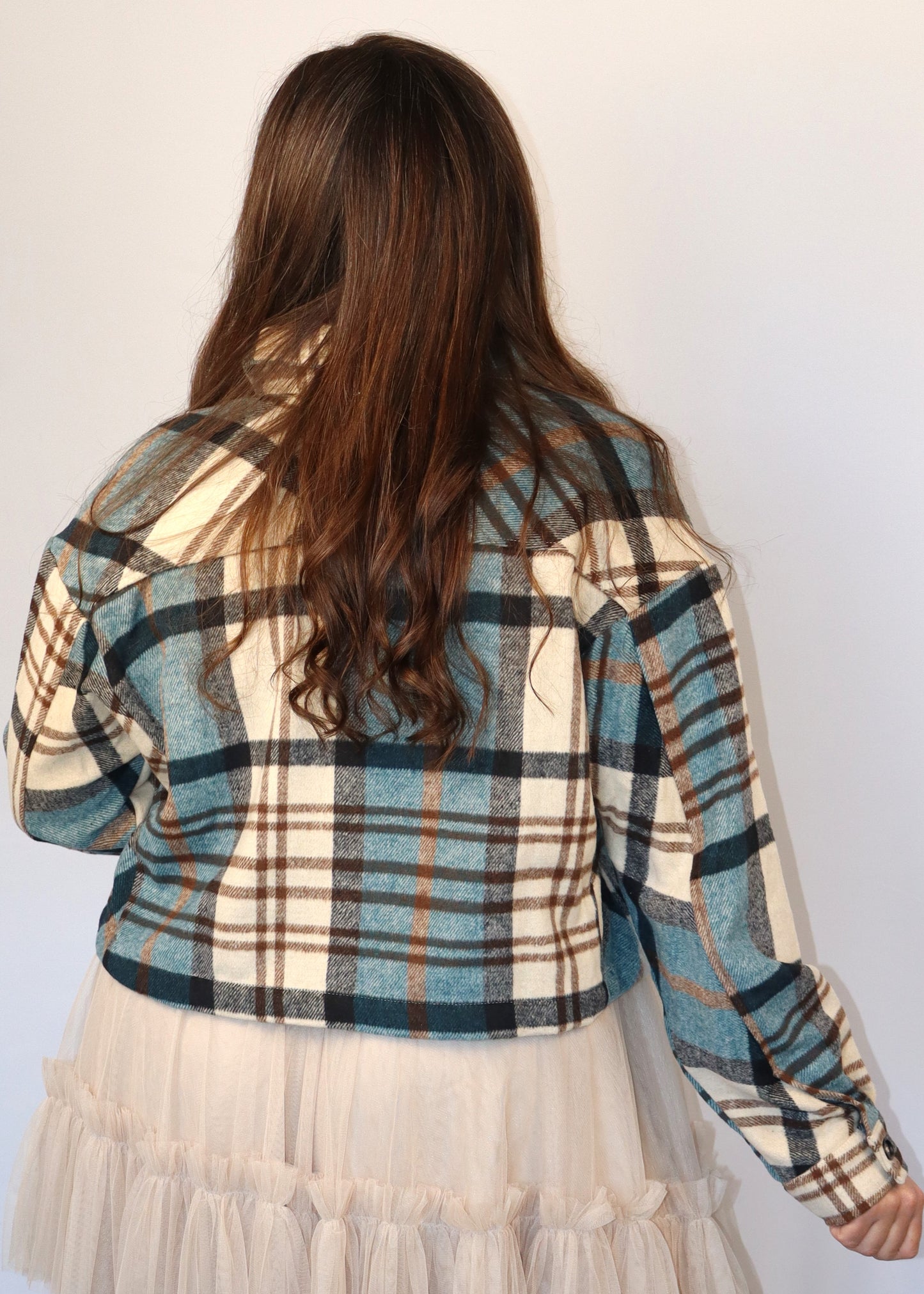 Woman wearing a teal, brown, and black cropped flannel jacket with a white tank top, taupe tiered tulle skirt, and multi-strand pearl necklace against a plain background
