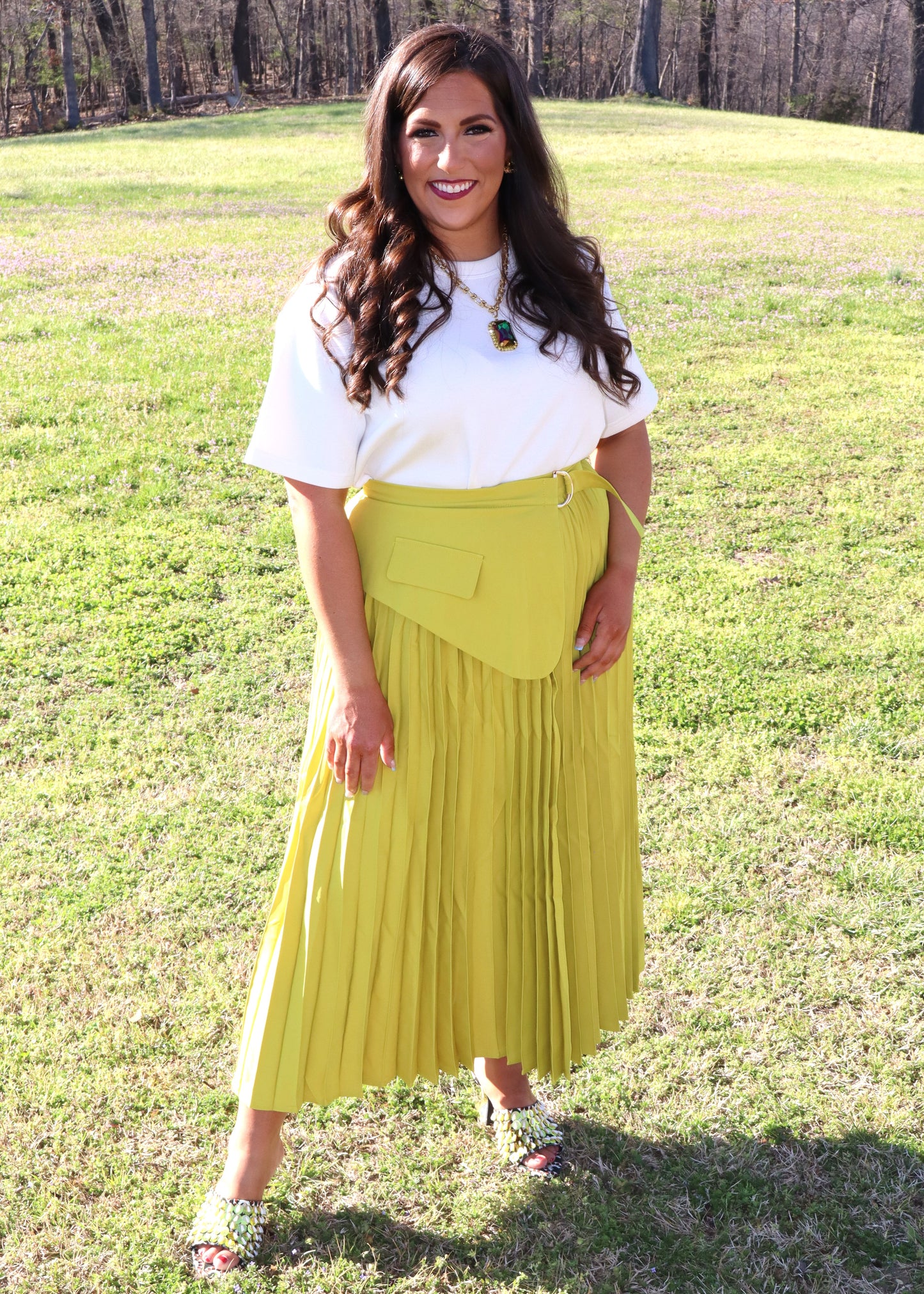 Woman wearing a white top and yellow pleated skirt standing in a grassy field.