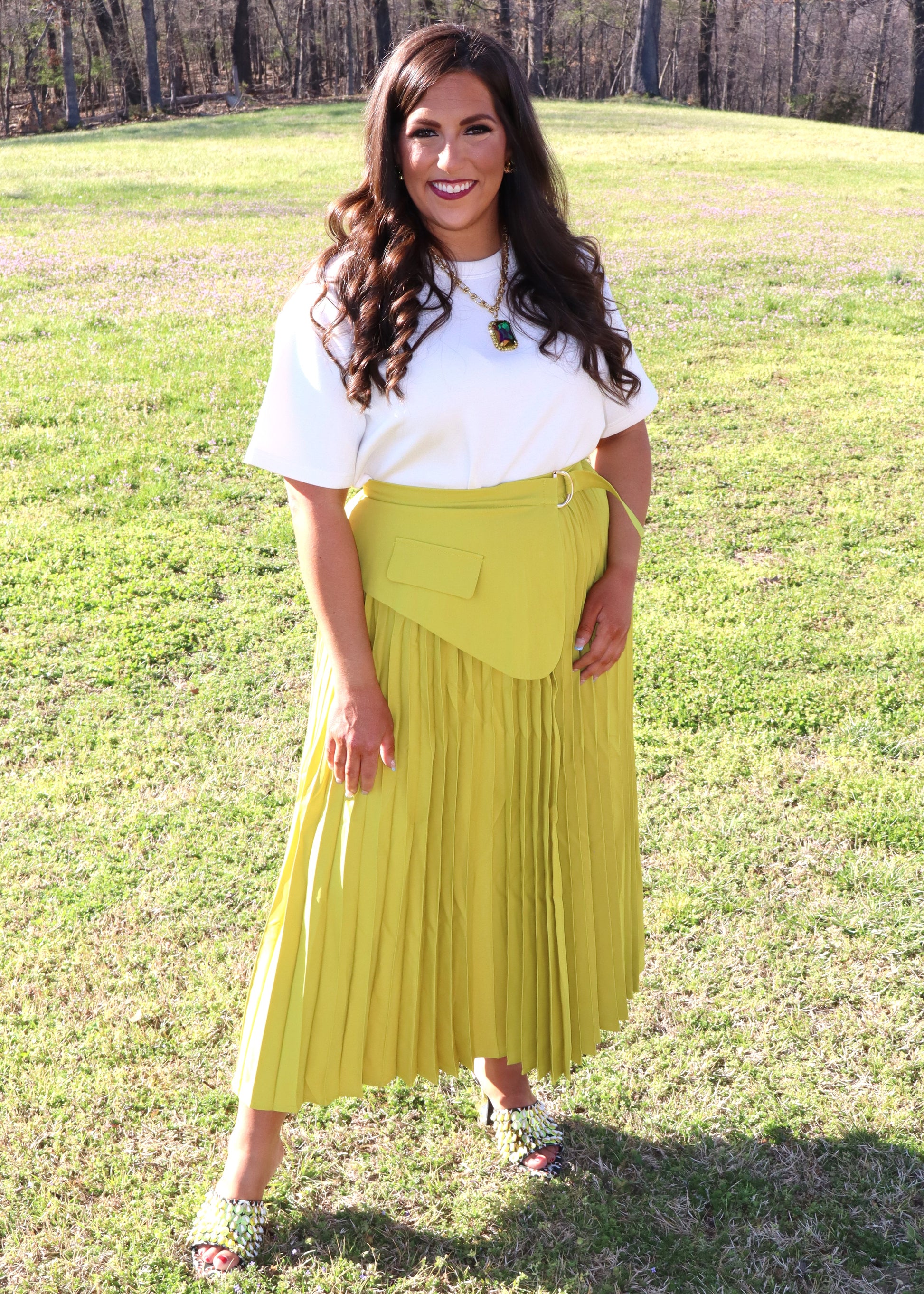 Woman wearing a white top and yellow pleated skirt standing in a grassy field.