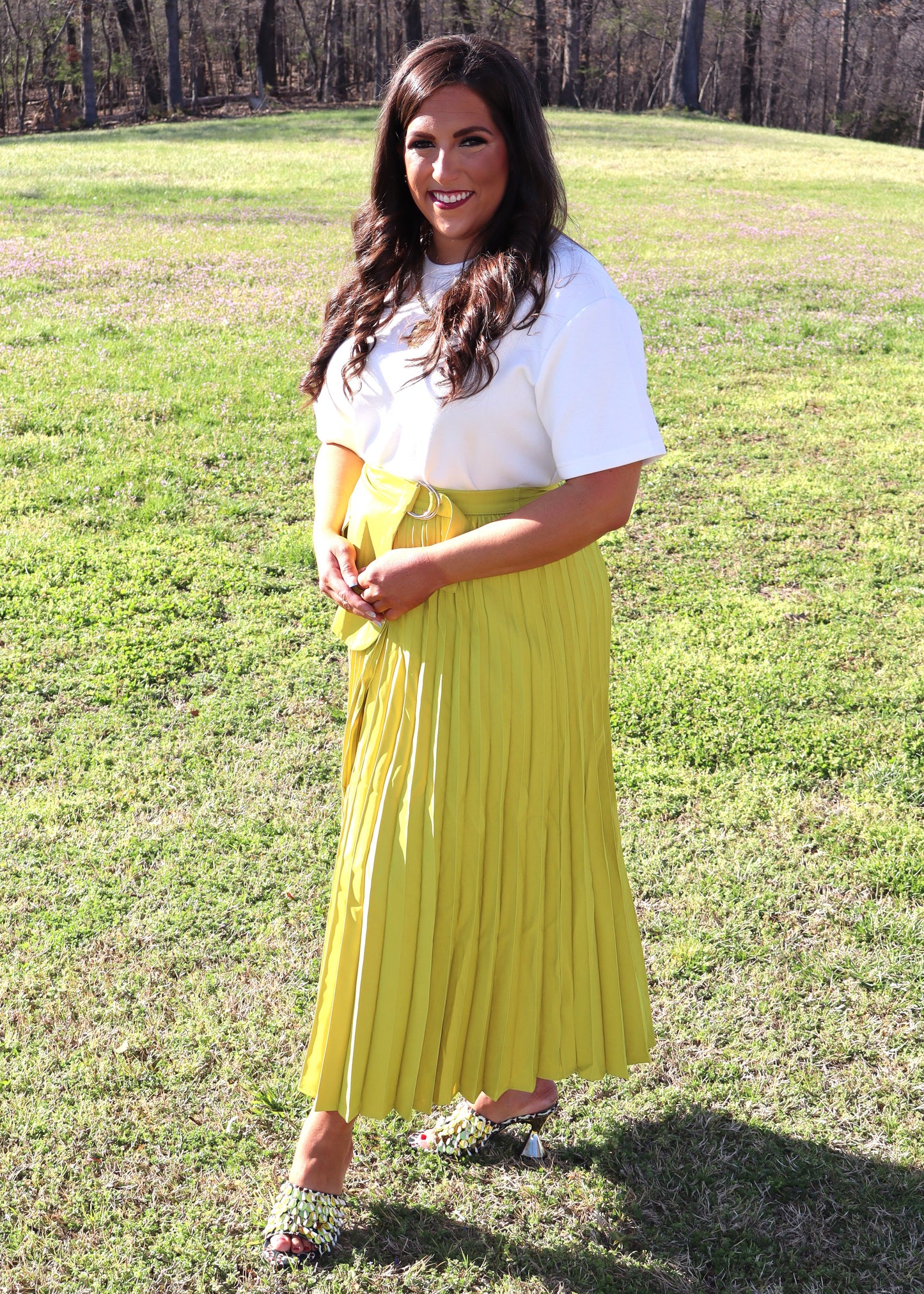 Woman wearing a white top and yellow pleated skirt standing in a grassy field.