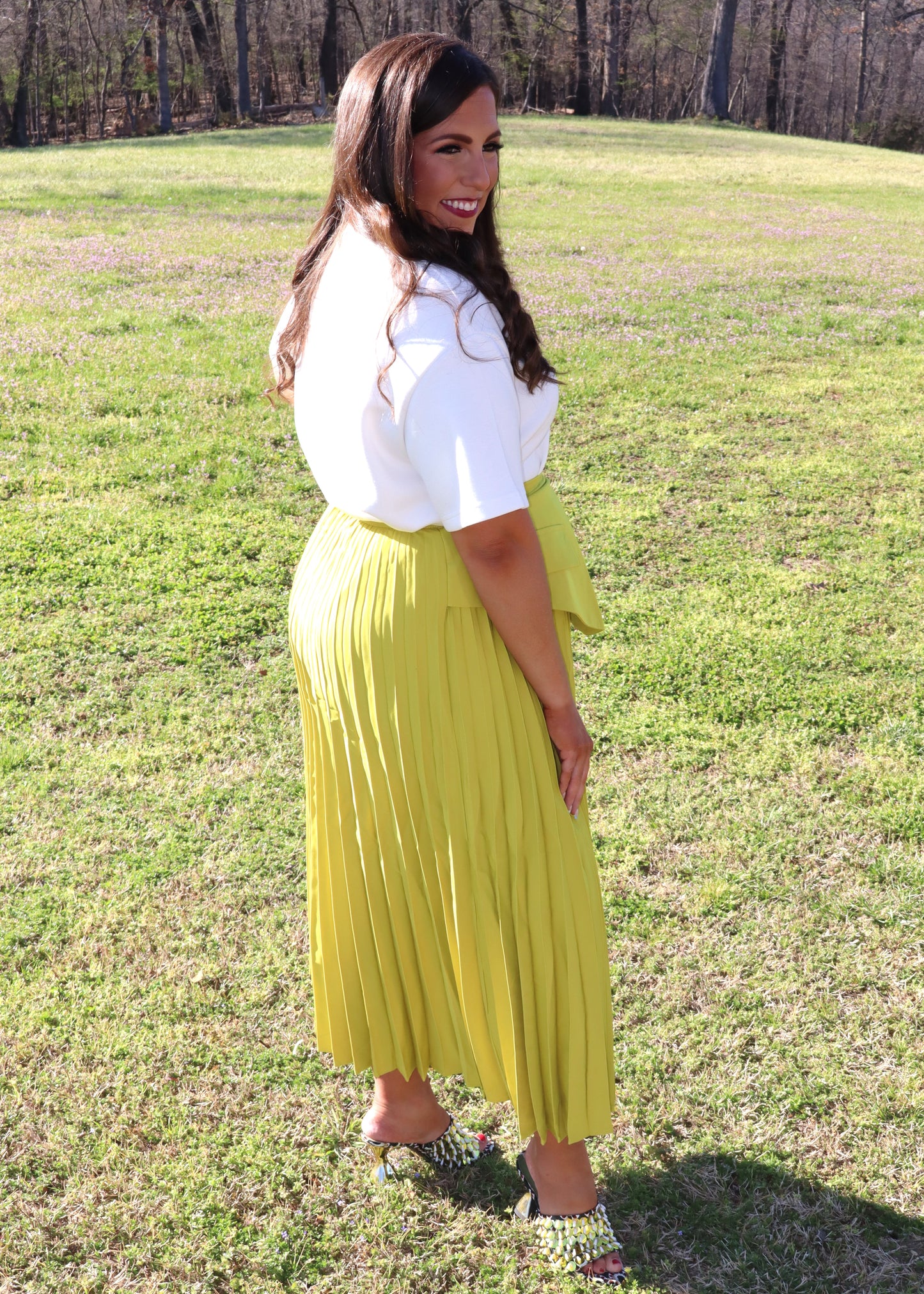 Woman in a white top and yellow pleated skirt standing in a grassy field.