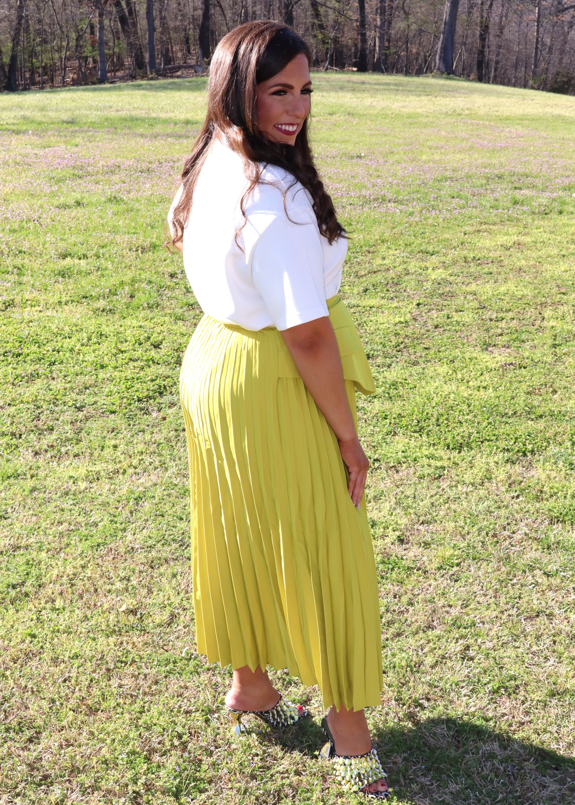 Woman in a white top and yellow pleated skirt standing in a grassy field.