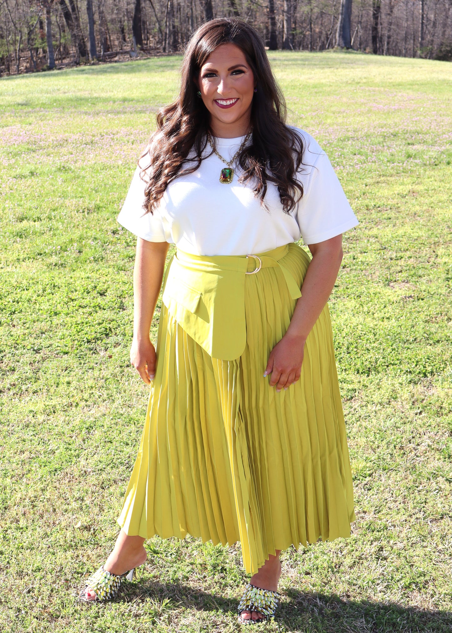 Woman wearing a white top and yellow pleated skirt standing in a grassy field.