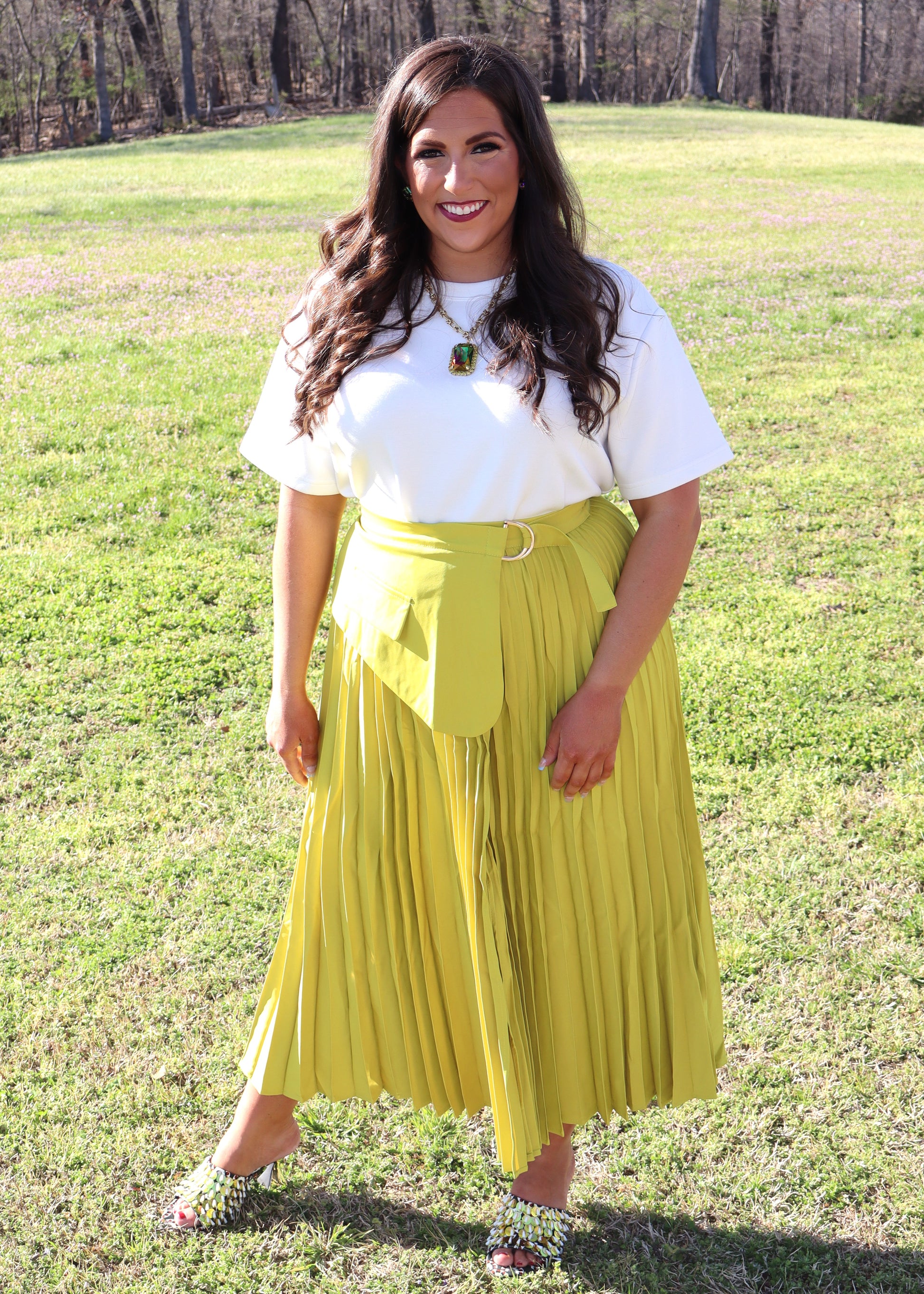 Woman wearing a white top and yellow pleated skirt standing in a grassy field.